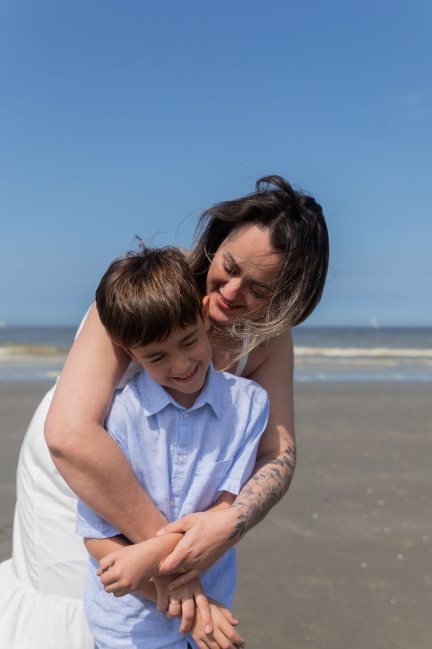 A smiling mother hugs her young son from behind on a sunny beach with ocean waves. photography brussels