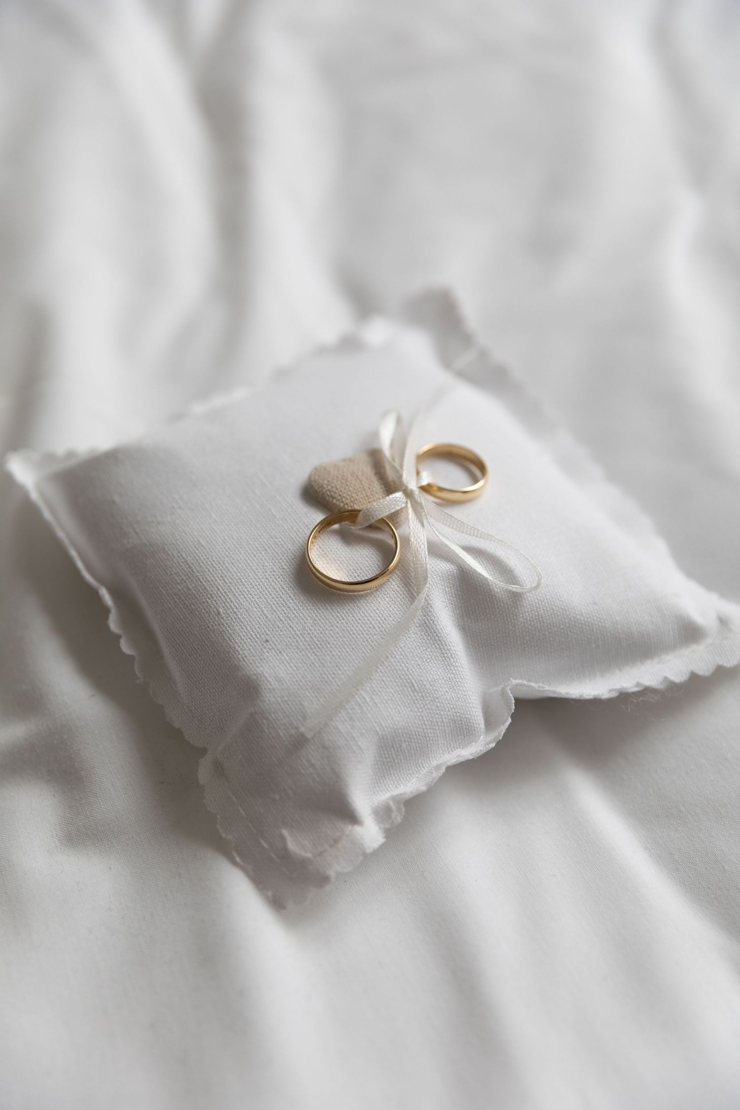 Two gold wedding bands resting on white lace ring pillow tied with ribbon on white fabric.