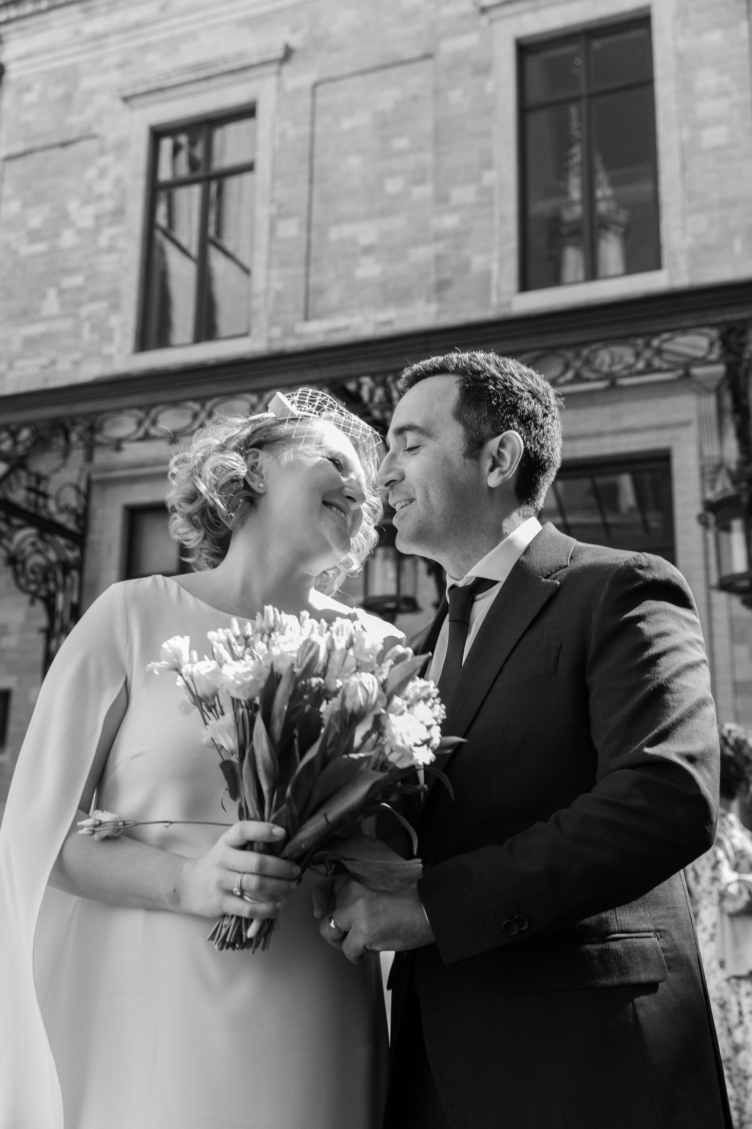 Black and white photo of a bride and groom sharing a romantic moment, bride holding tulip bouquet outside historic building.