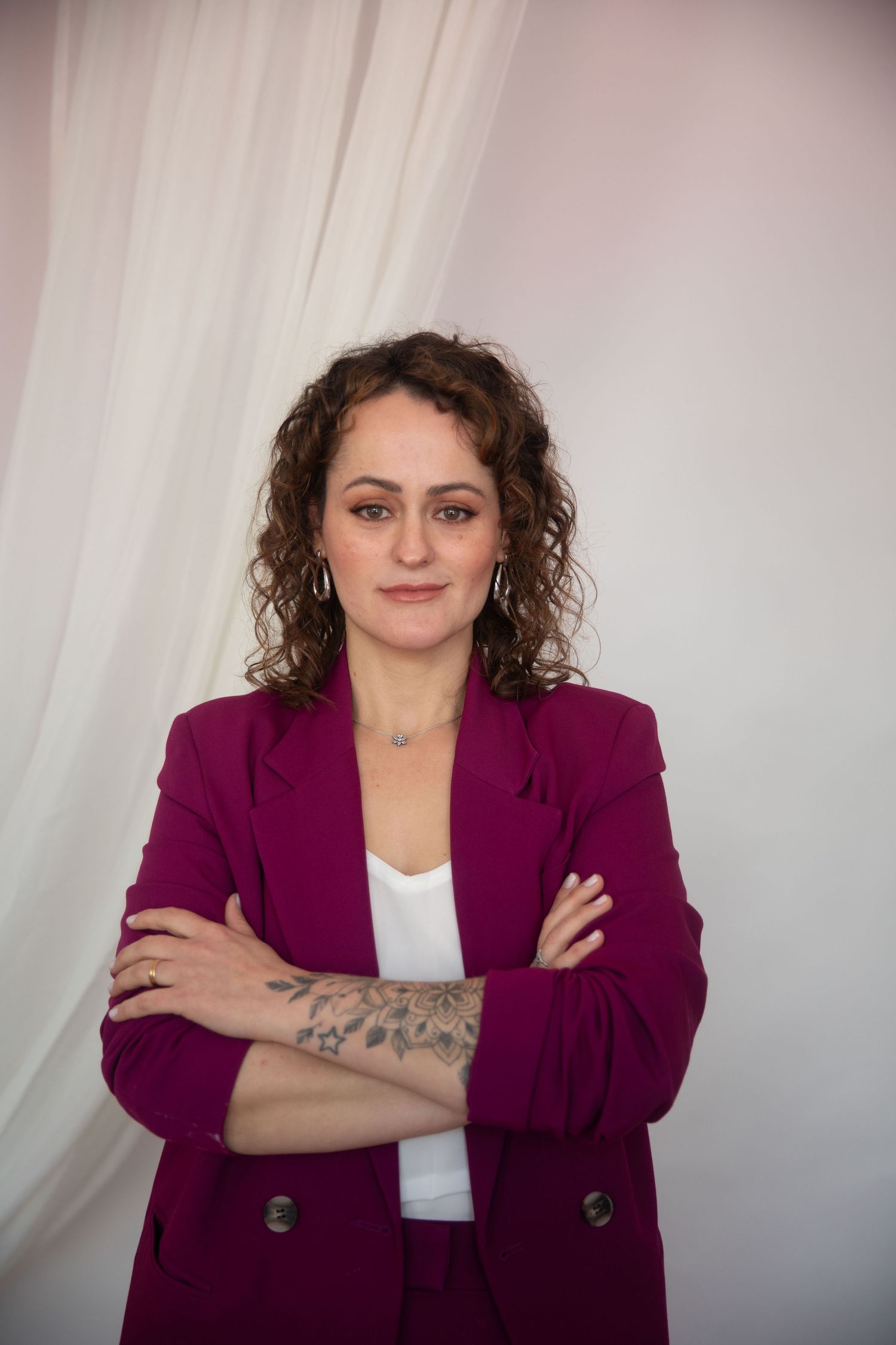 Woman in magenta blazer with tattoos, arms crossed, curly hair, posing confidently against white draped background.