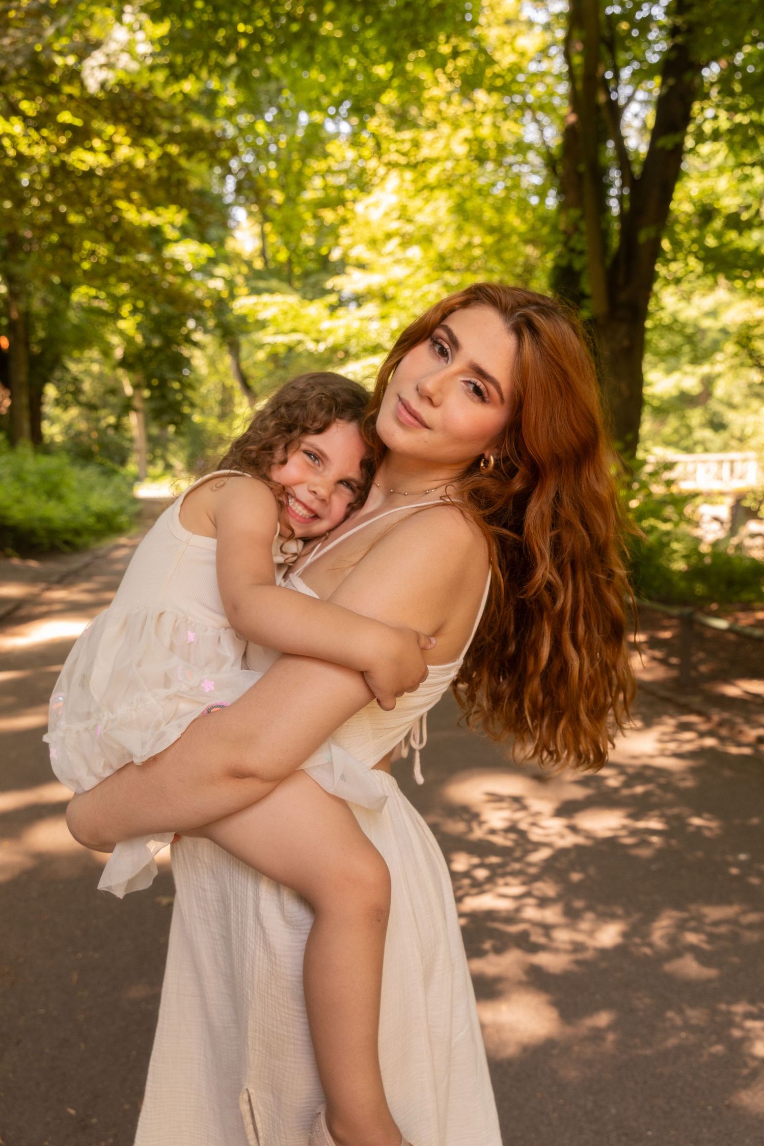 A young woman with red hair holds a smiling child in a sunny park, both wearing white dresses.