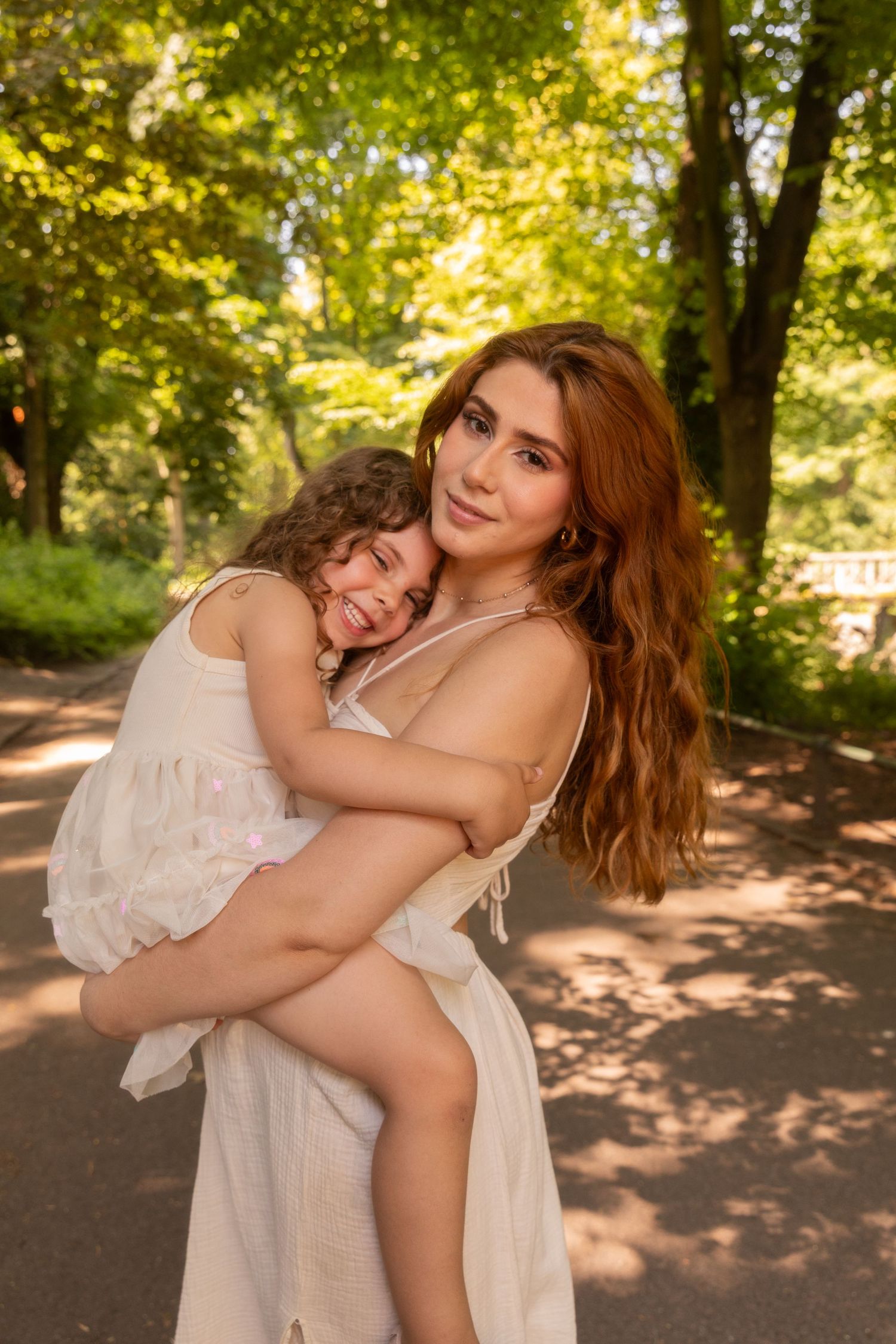 A woman with long red hair holds a young girl in a sunny park, both wearing white dresses and smiling warmly.