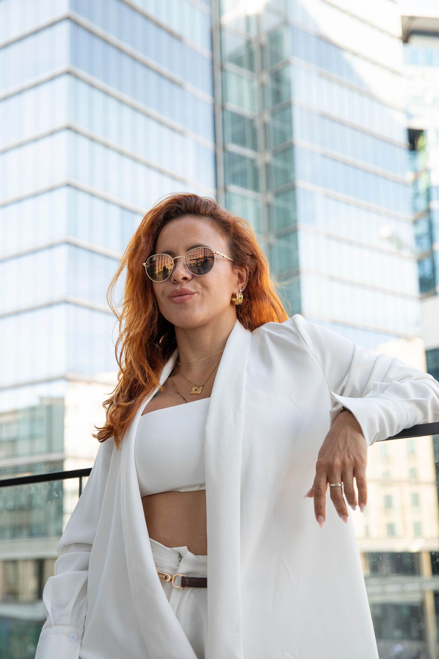 Stylish woman with red hair in white blazer and sunglasses posing against modern glass building exterior.