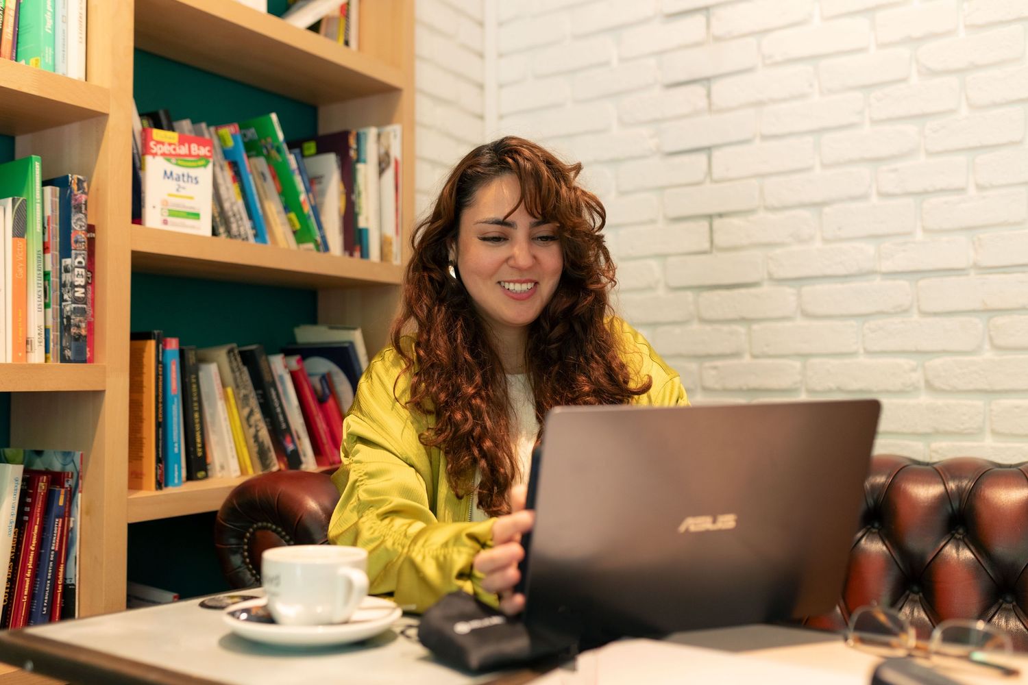 Young woman with curly hair in yellow jacket smiling at ASUS laptop in cozy cafe with bookshelves.