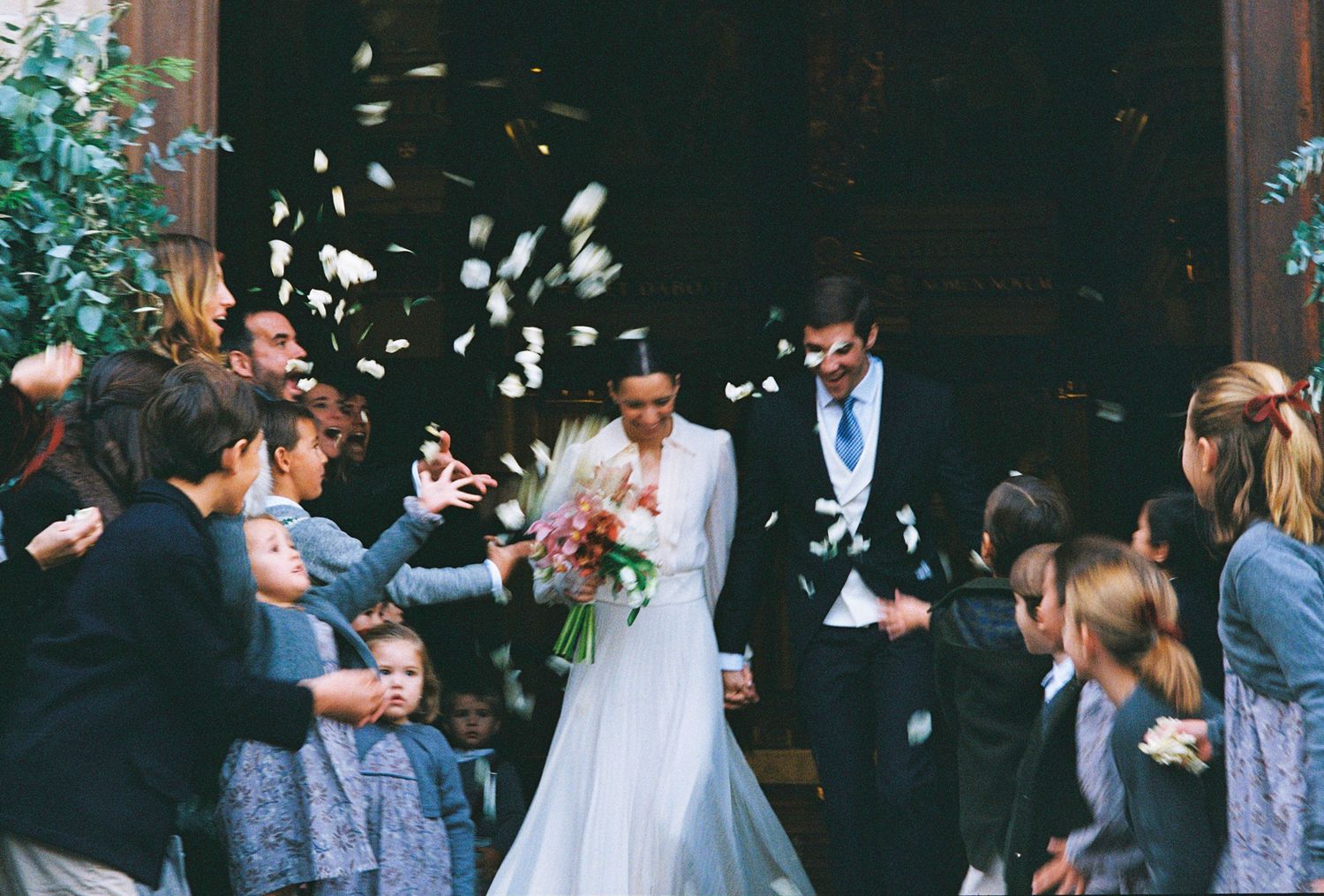 Bride and groom exit church as guests toss white flower petals in joyful wedding celebration.