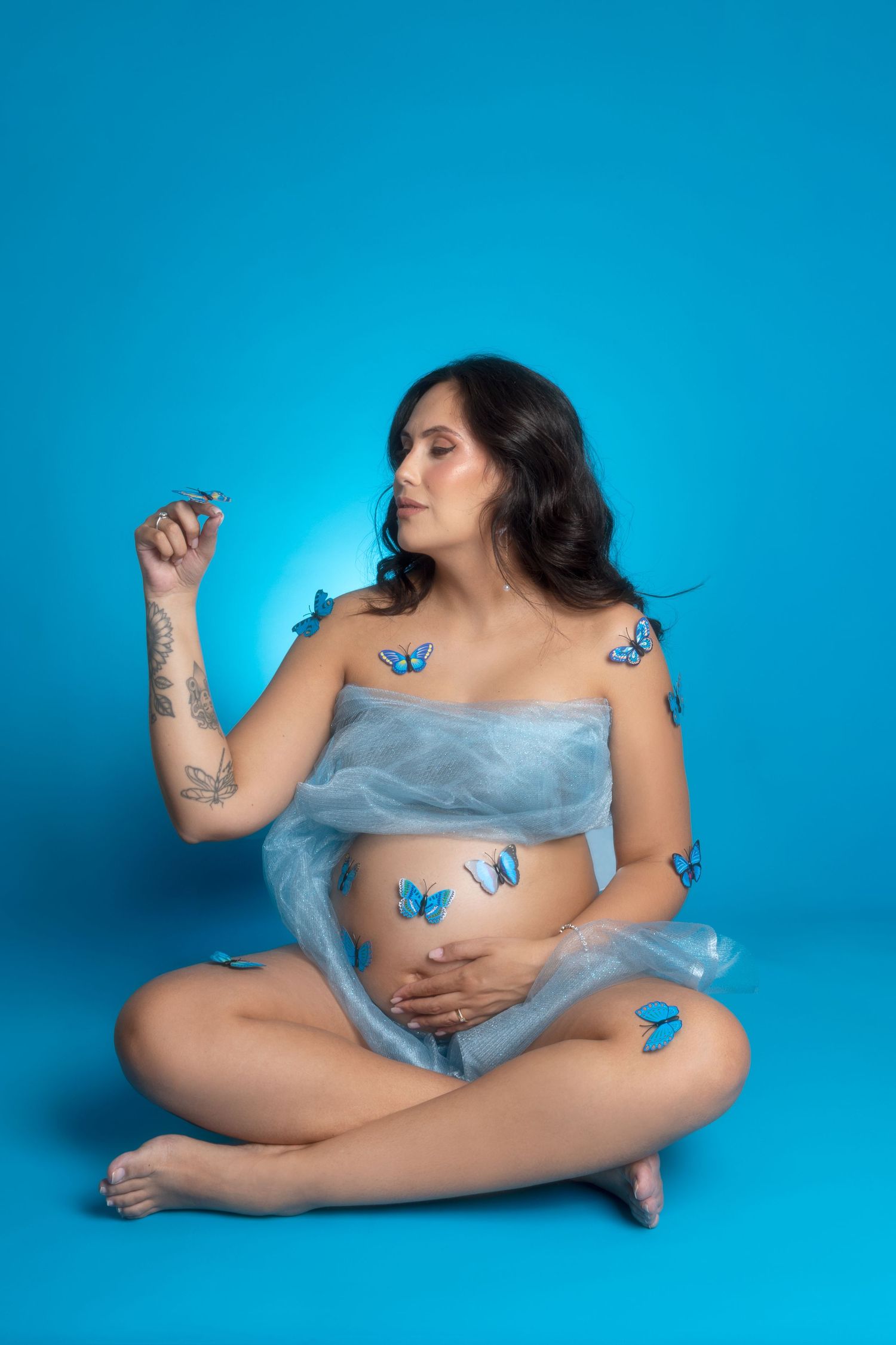 Pregnant woman sitting cross-legged with blue butterflies on her body, wrapped in sheer fabric against a blue background.