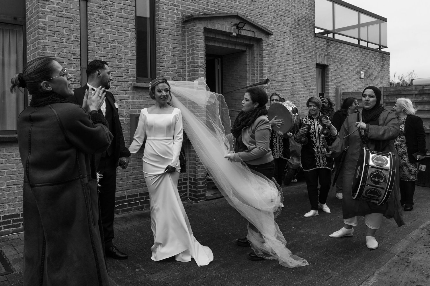 Black and white photo of a bride in a white gown and veil walking outside with wedding guests celebrating.