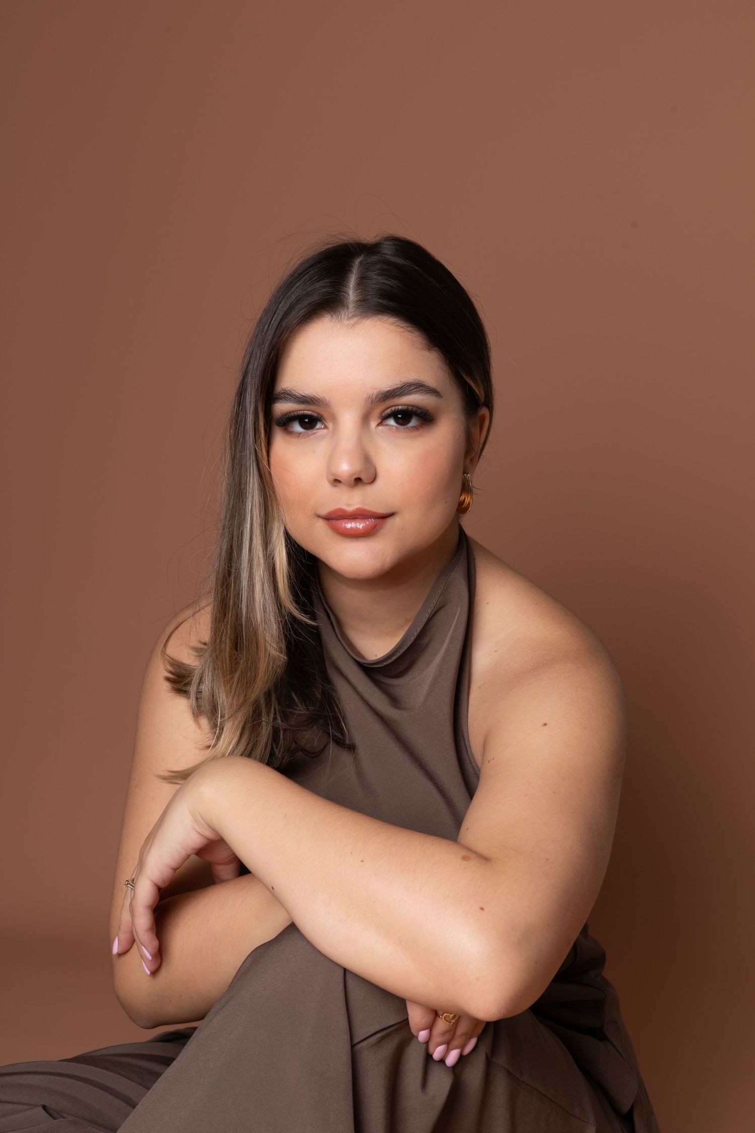 Young woman in brown sleeveless dress sits posed with arms crossed, wearing gold earrings against a warm brown background.