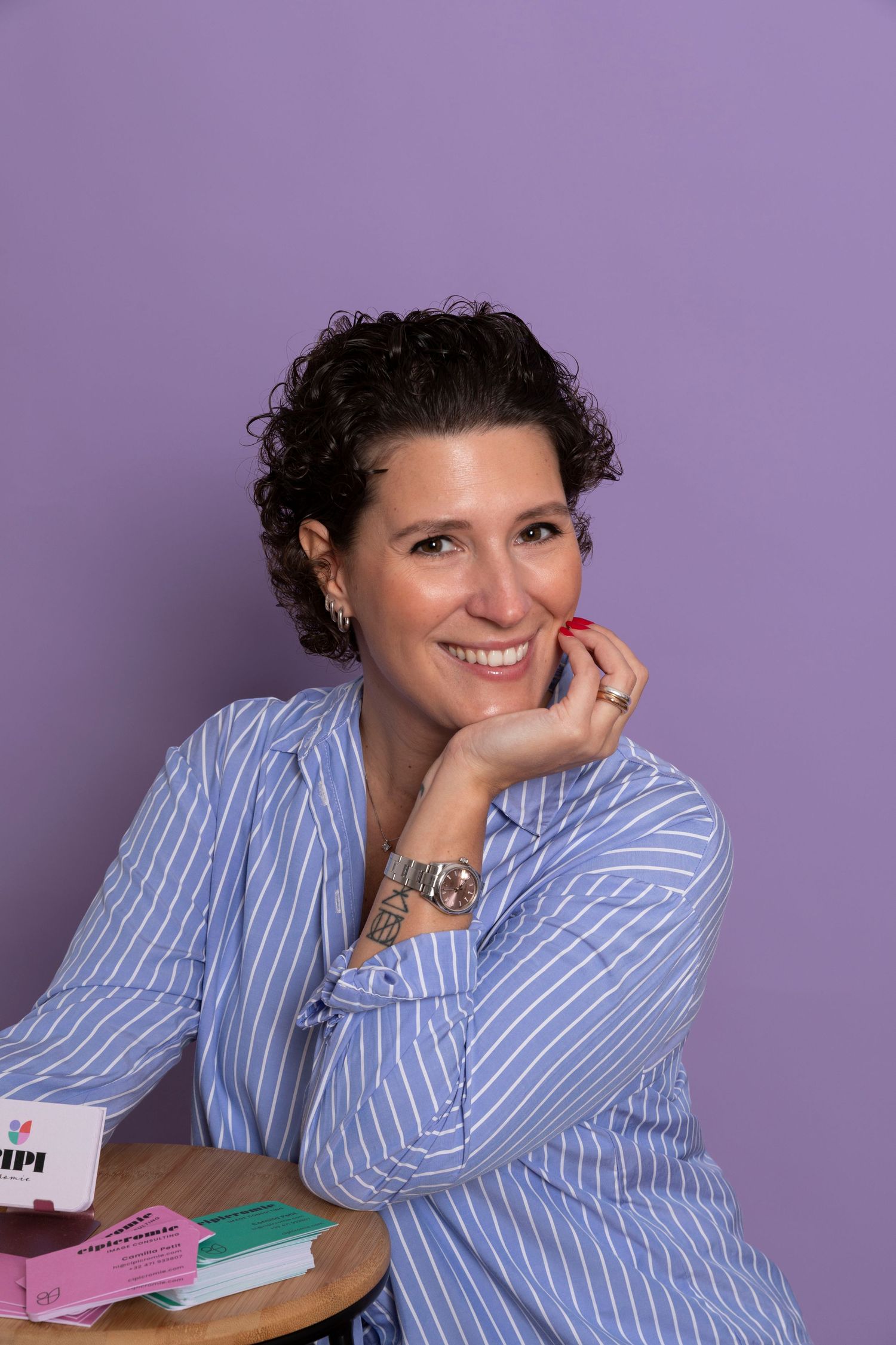 Smiling woman in blue striped shirt sits at table with books, purple background, tattoo visible on wrist.