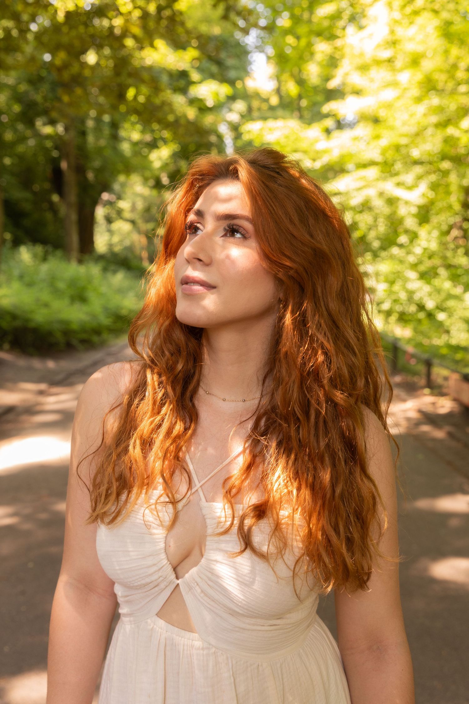 Young woman with long red wavy hair in a white dress gazing upward in a sunlit green park.