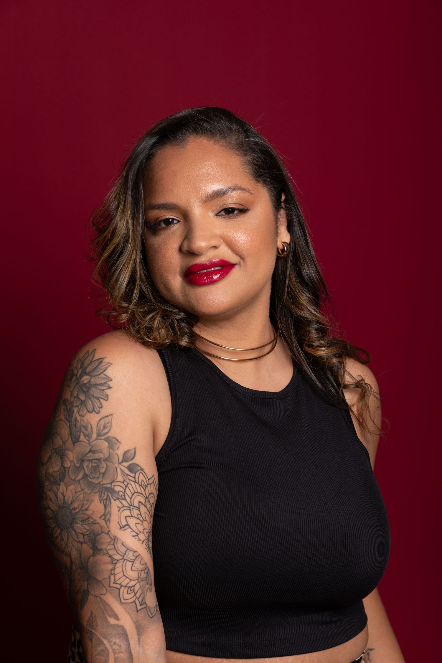 Woman with floral arm tattoos in black crop top poses against red background with bold red lips and highlighted hair.