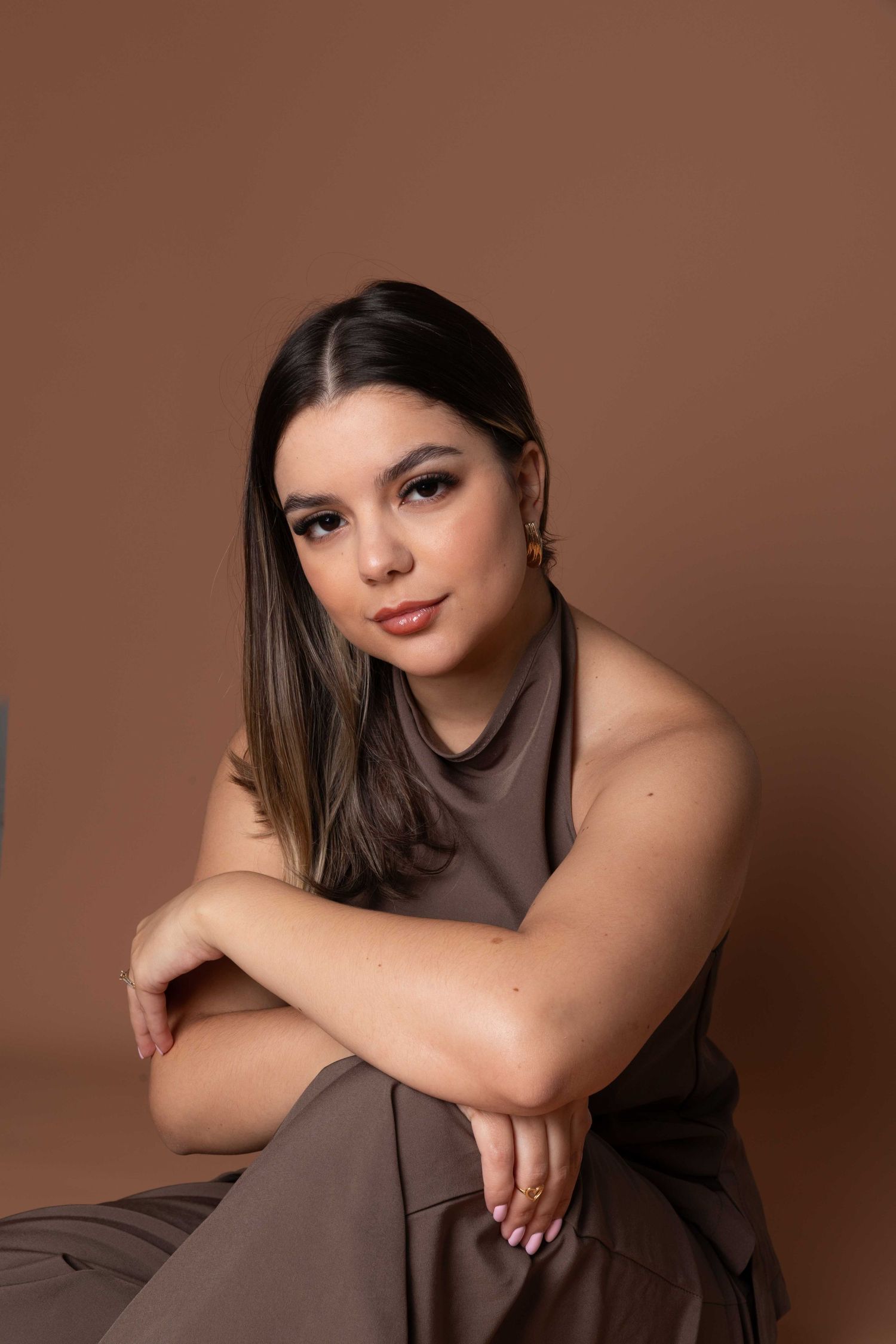 Young woman in brown outfit sits gracefully against a warm beige backdrop, wearing gold earrings and pink nails.