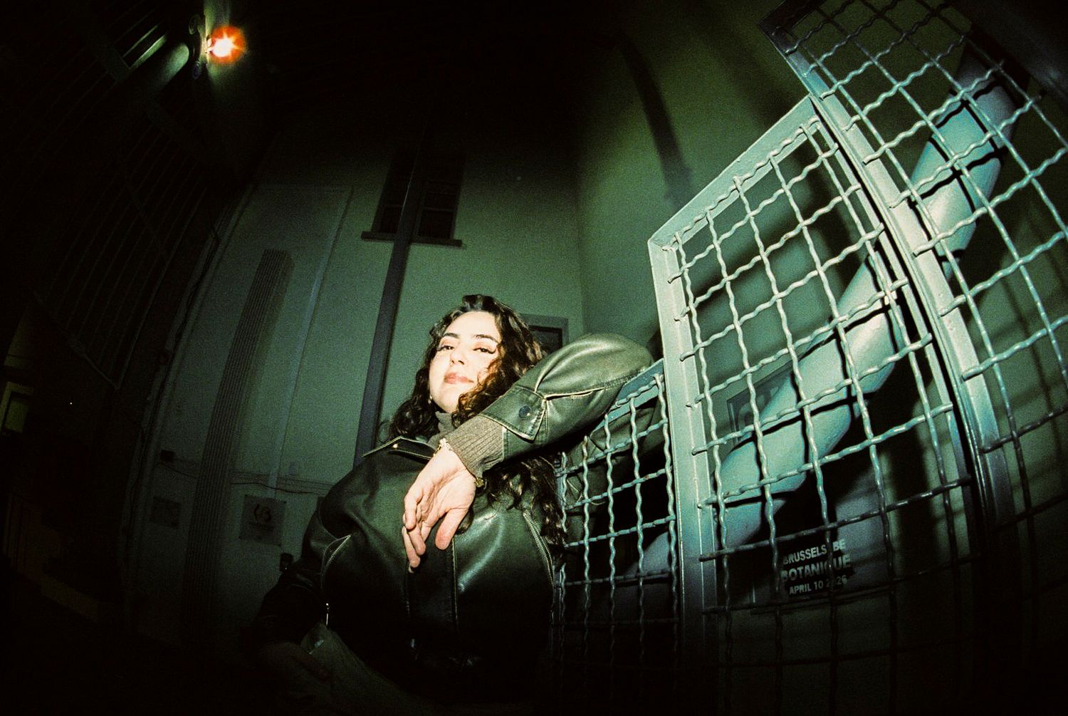 Woman in black leather jacket leans against a metal gate in a moody, dimly lit setting. photography brussels