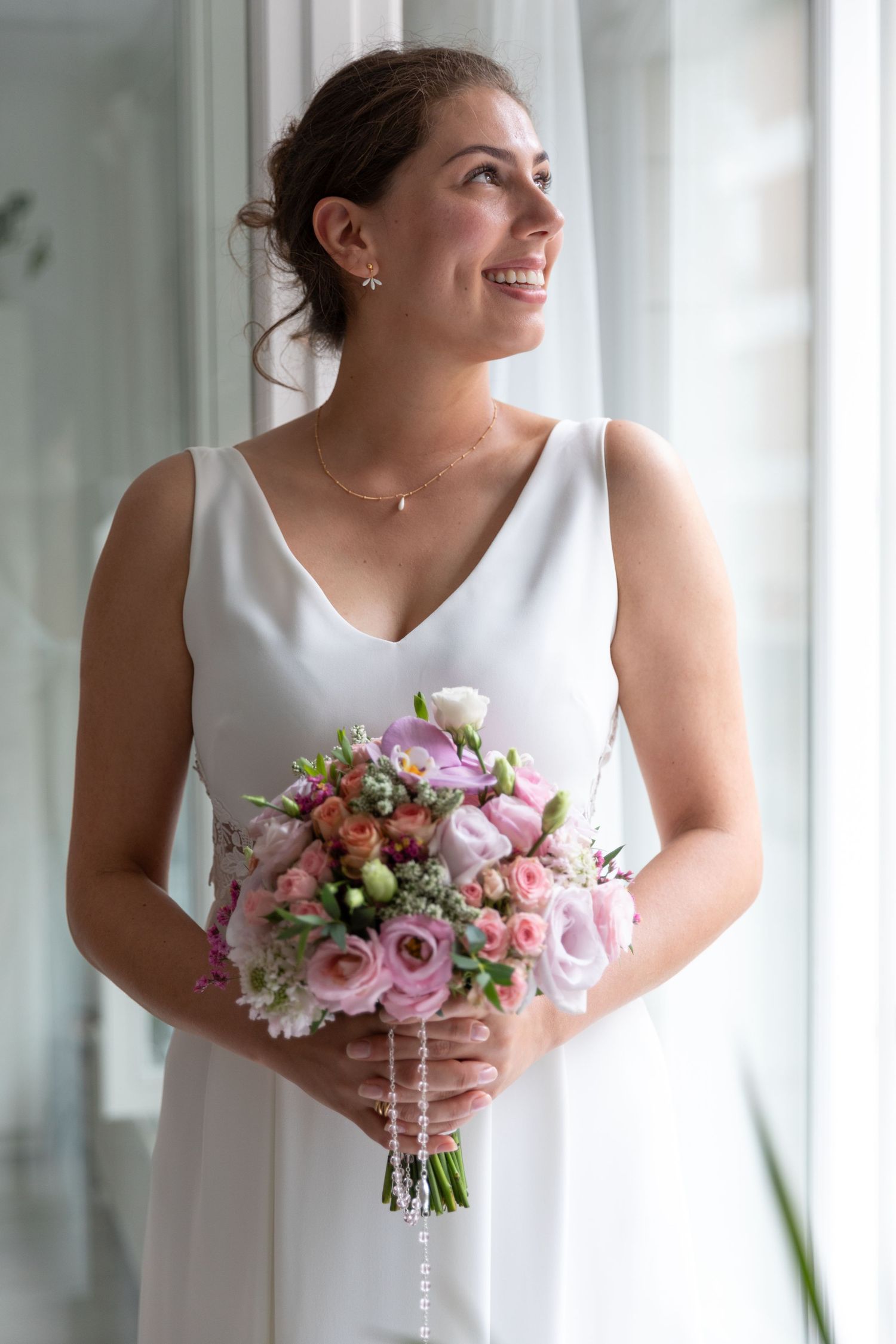 Smiling bride in white sleeveless gown holding colorful pink and purple floral bouquet near window.