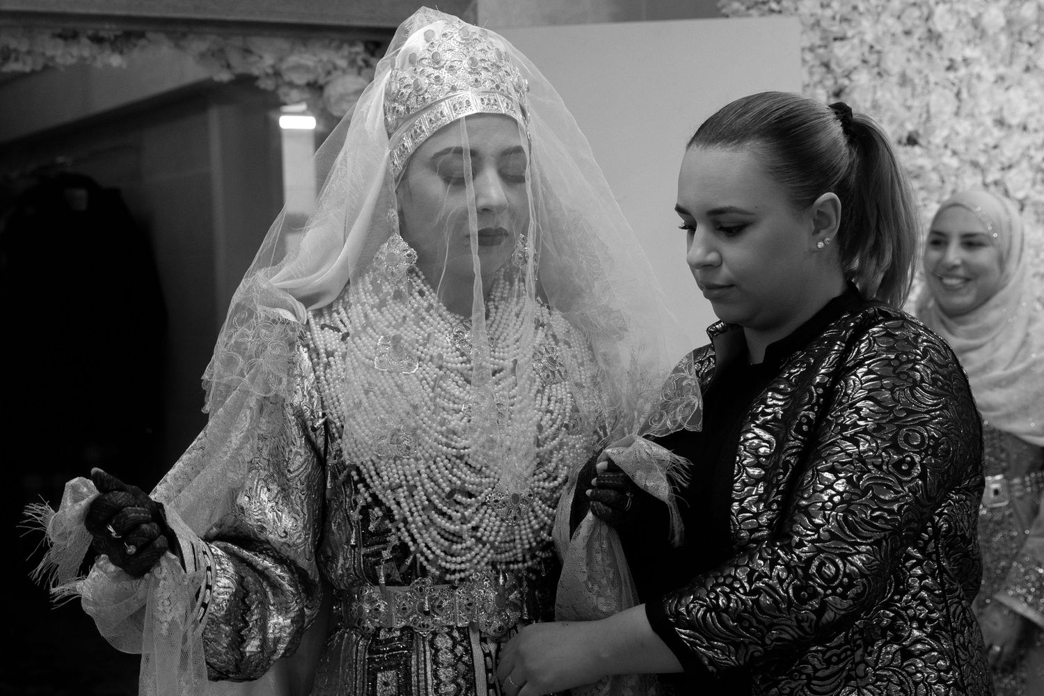 Black and white photo of a bride in ornate lace wedding dress and veil being assisted by a woman.