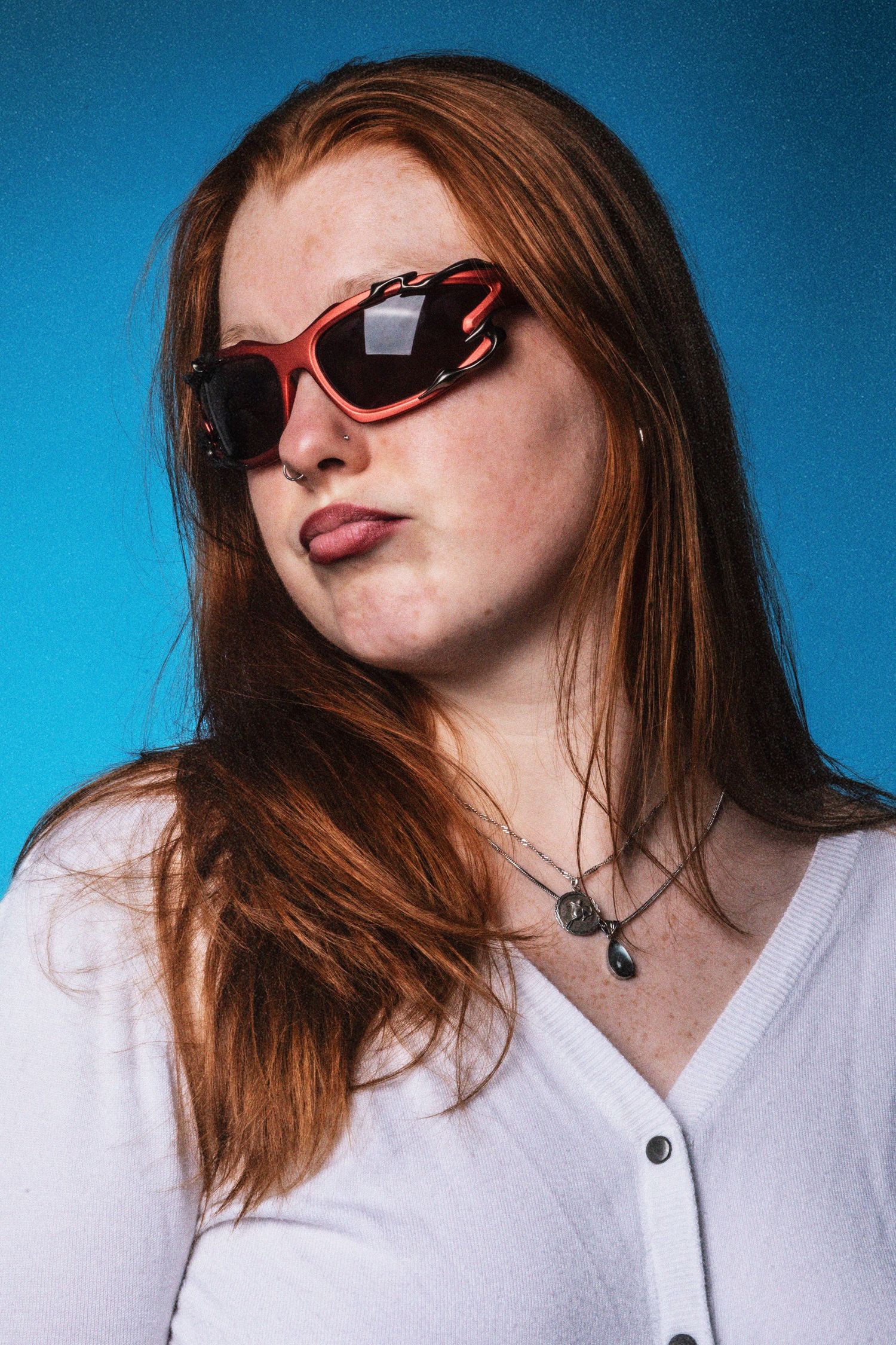 Young redhead woman wearing red heart-shaped sunglasses and white cardigan poses against blue background.