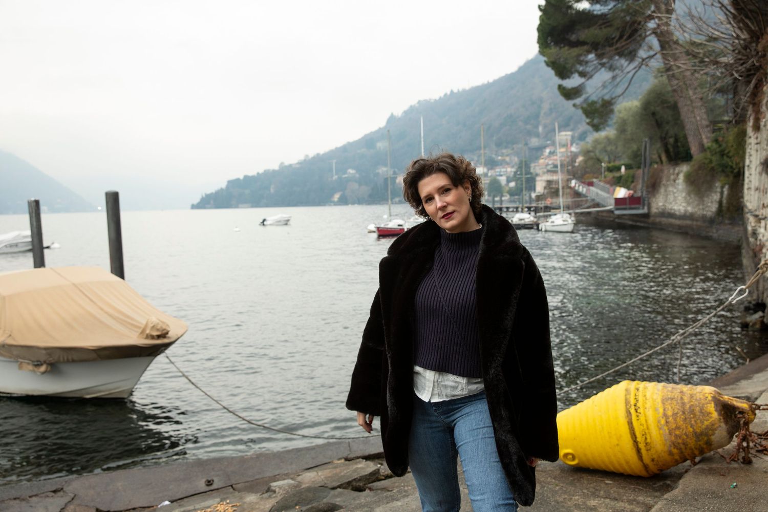 Woman in black coat standing by a lakeside dock with moored boats and misty mountain backdrop.
