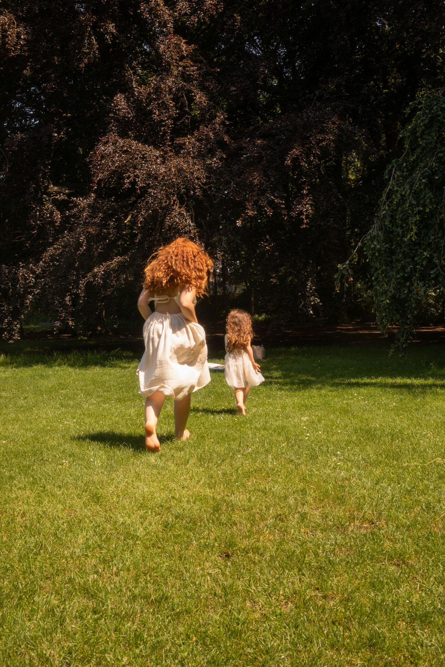 A redheaded mother and young daughter in matching white dresses run barefoot across green grass in a sunlit park.