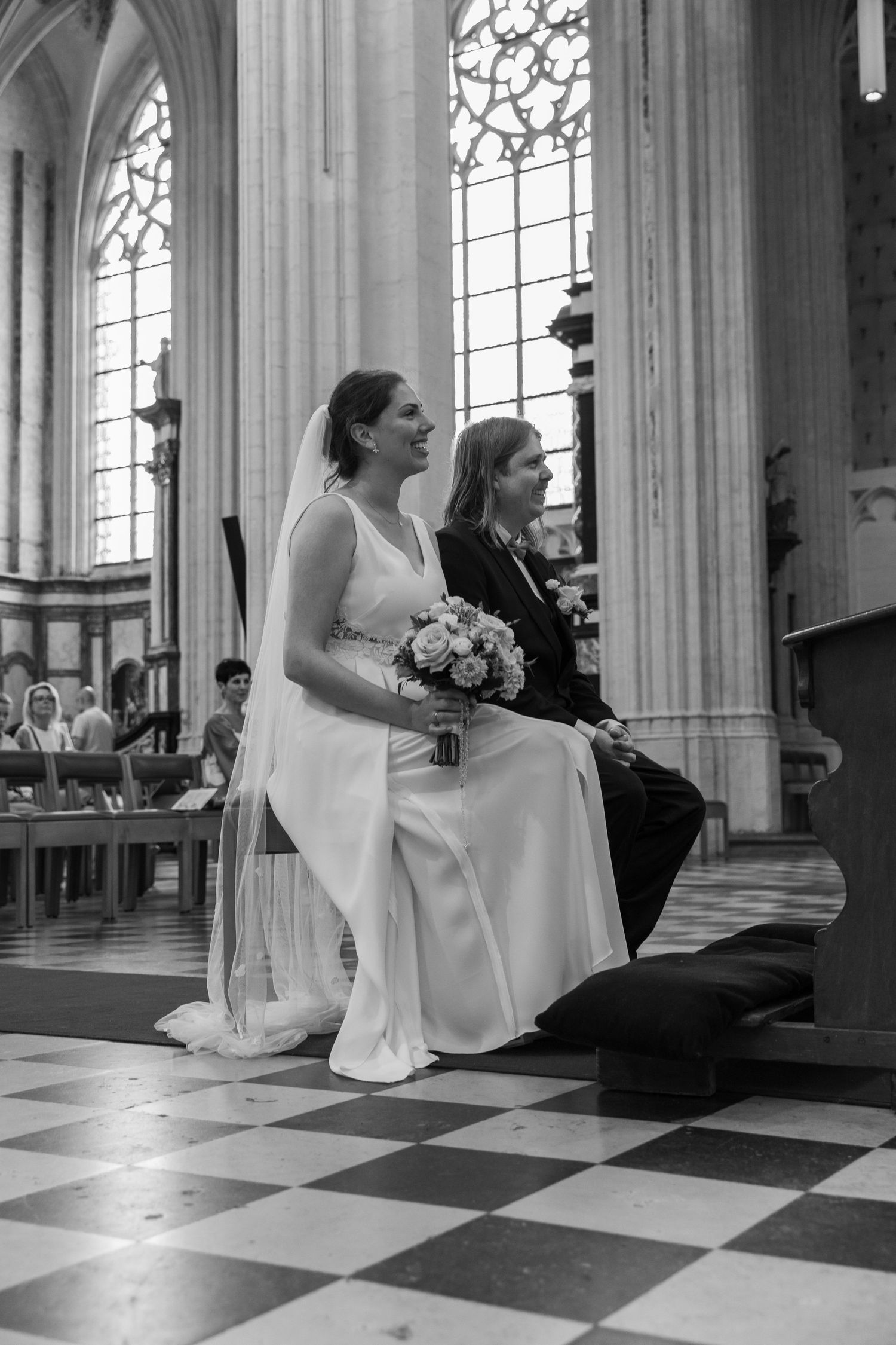 Bride in white gown holding bouquet sits with companion in ornate church during wedding ceremony.