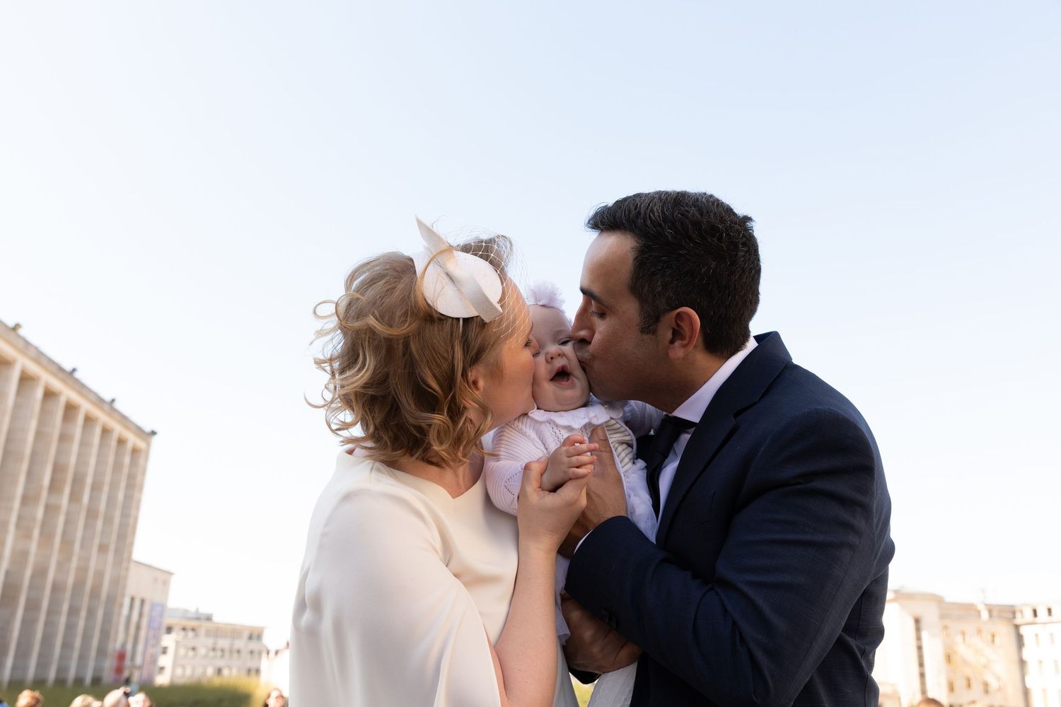 Happy parents kissing their laughing baby at an outdoor christening ceremony, father in navy suit.