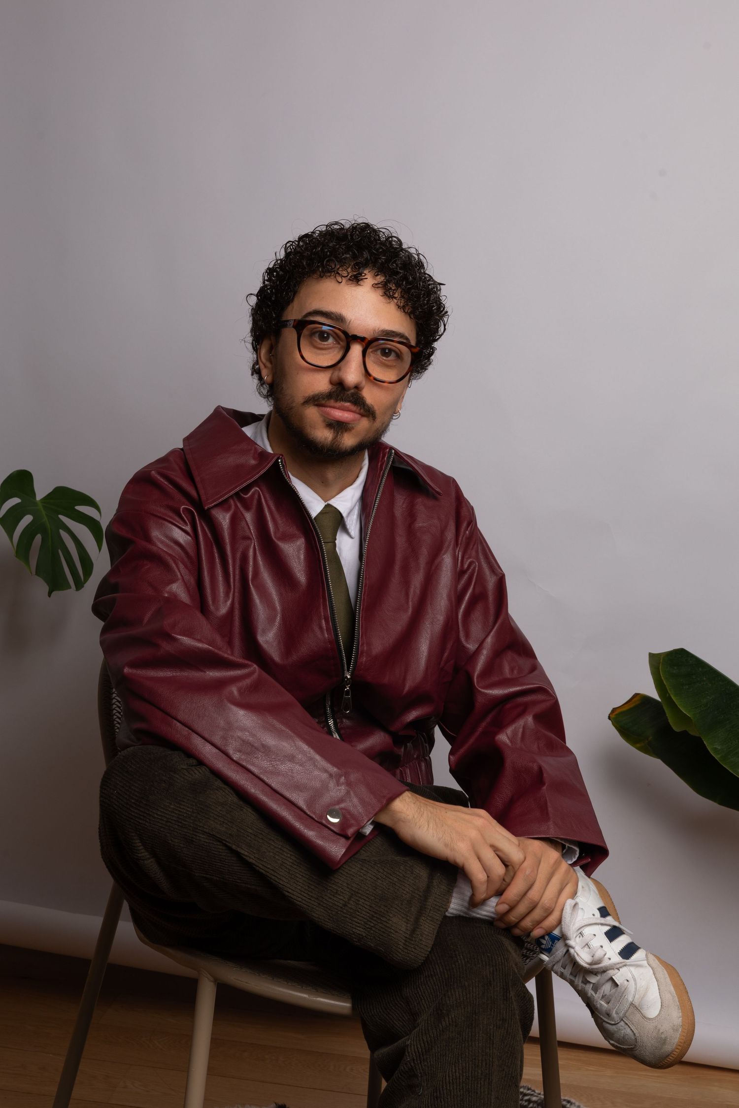 Man in burgundy leather jacket and glasses sitting on chair with plants in background.