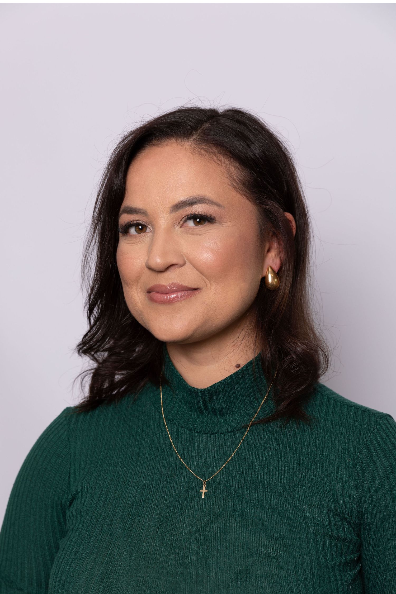 Woman in green turtleneck sweater with gold cross necklace and earrings, smiling against gray background.