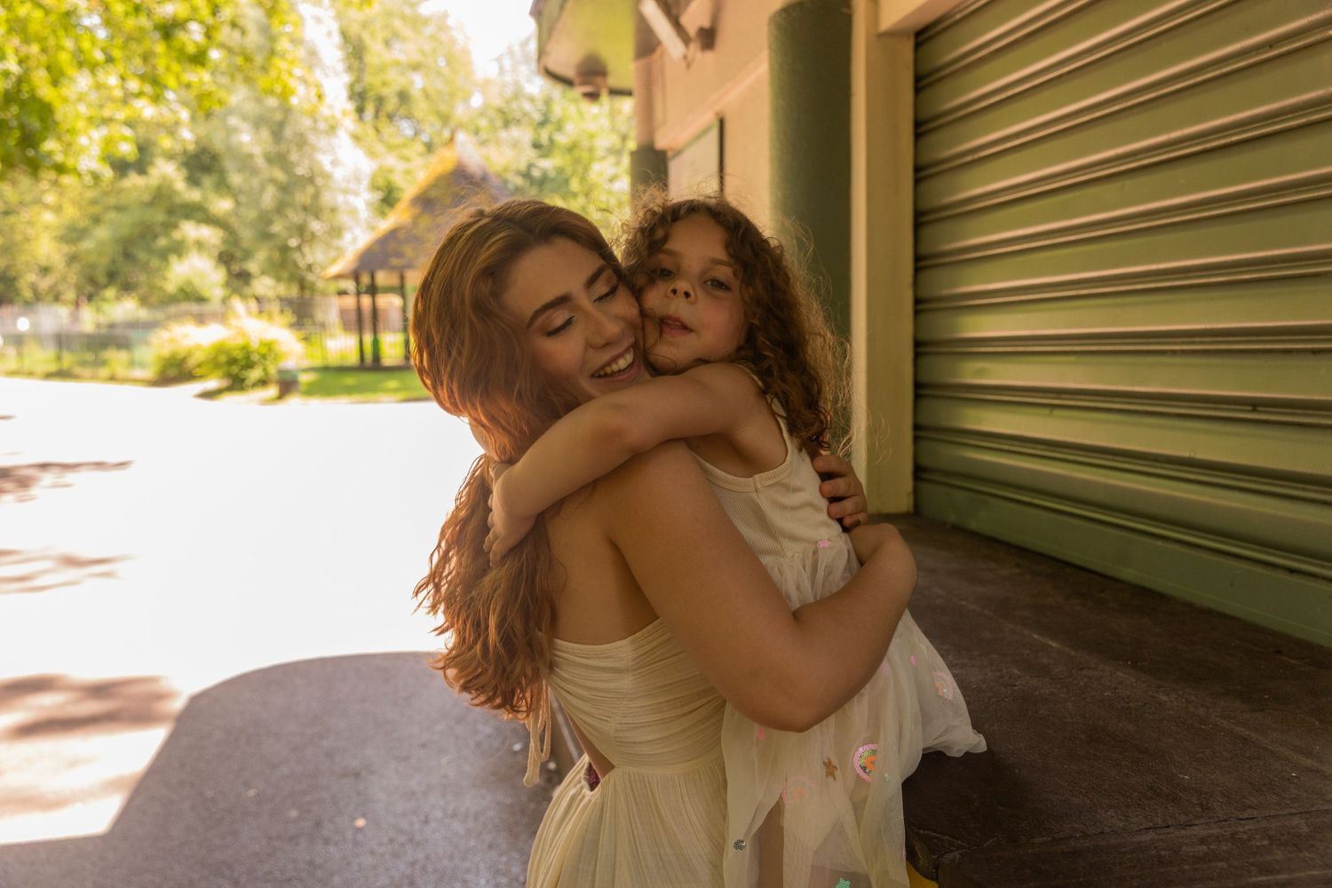 A mother in a cream dress tenderly holds and hugs her young daughter outdoors on a sunny day.