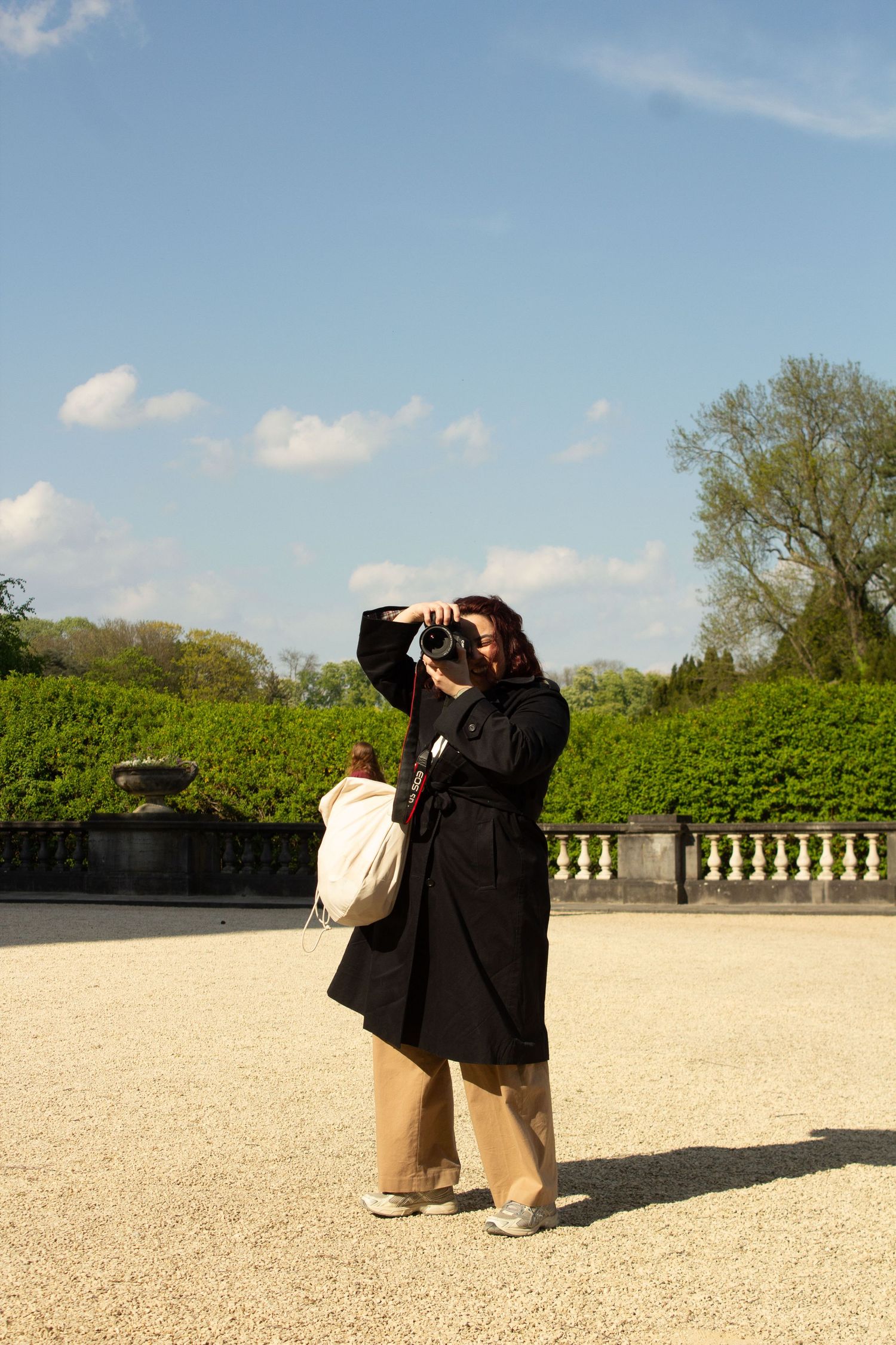 Woman in black coat photographs outdoors near manicured hedges and balustrade under blue sky.
