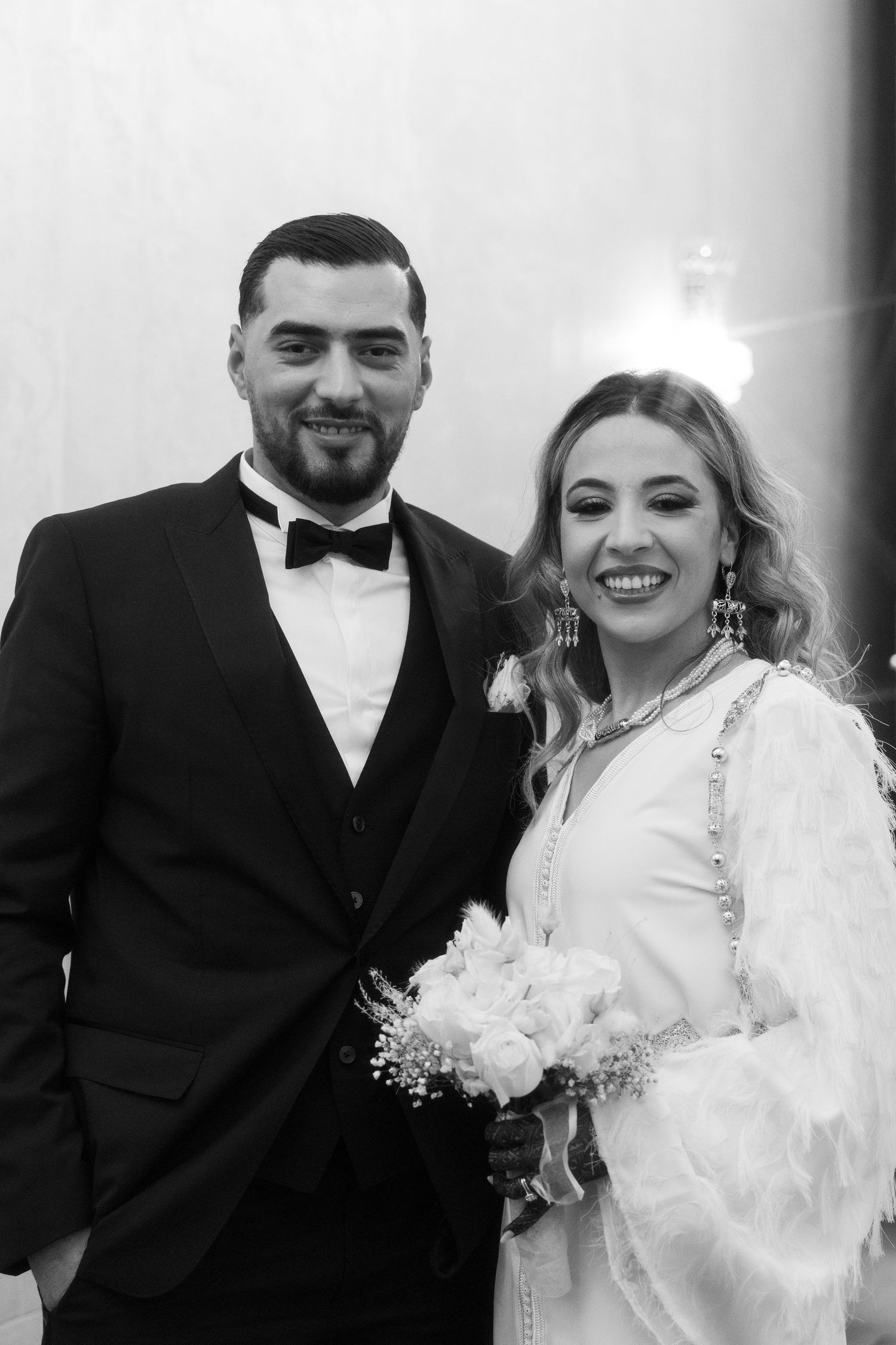Black and white wedding photo of smiling bride holding bouquet and groom in tuxedo posing together.
