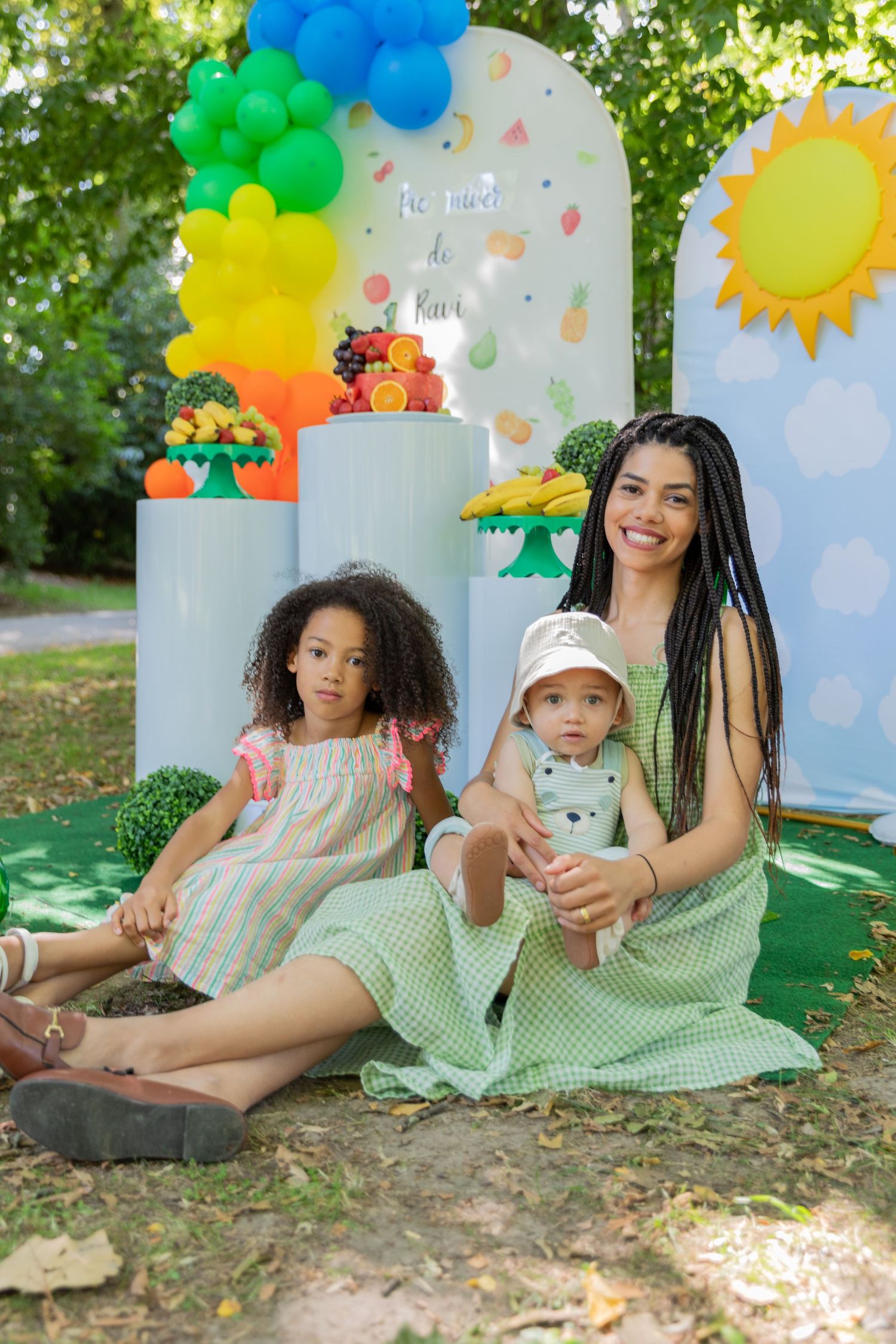 A woman with braids sits outdoors with two children at a colorful fruit-themed birthday party with balloon decorations.