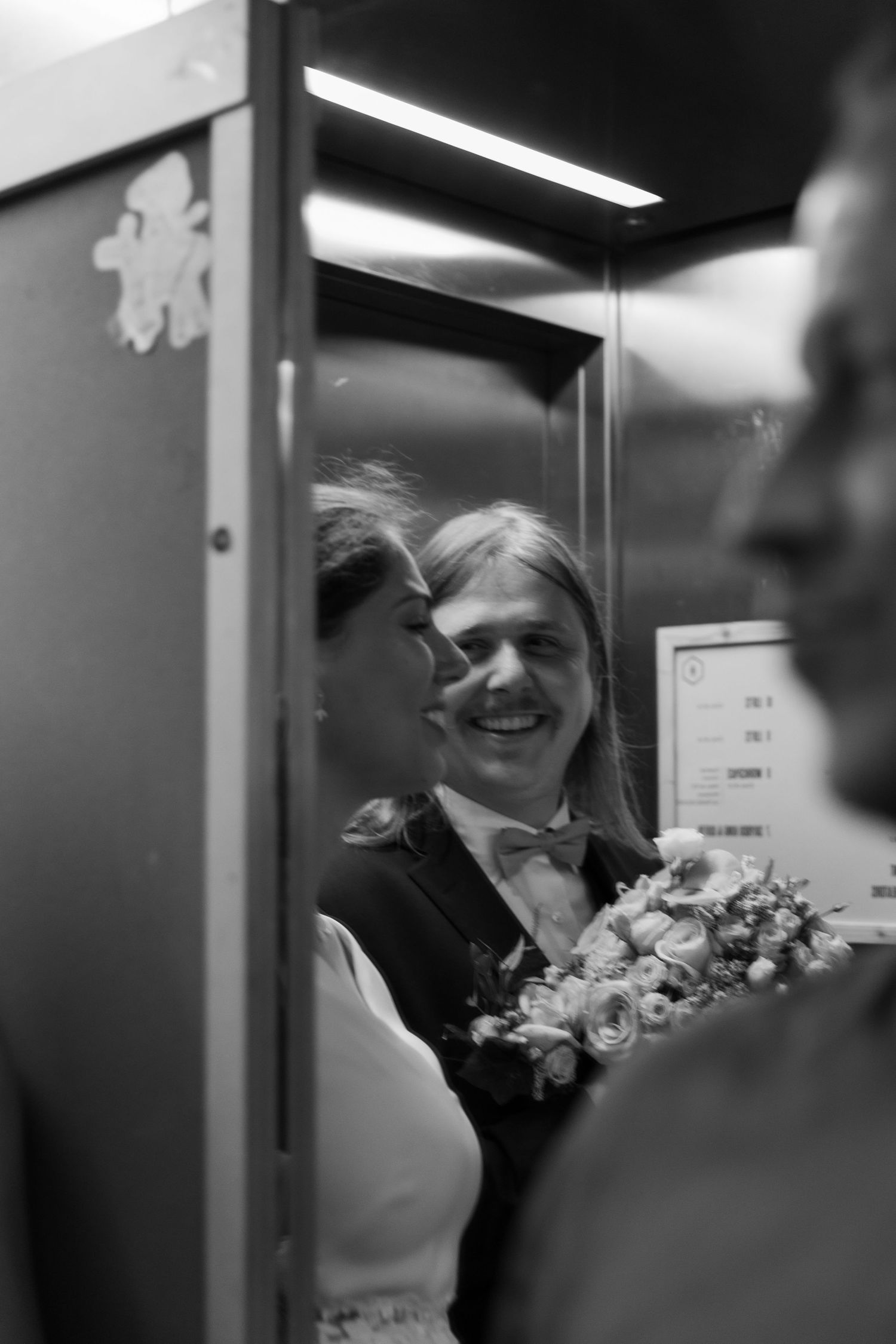 Smiling groom with long hair holds bouquet beside bride in elevator in black and white photo.