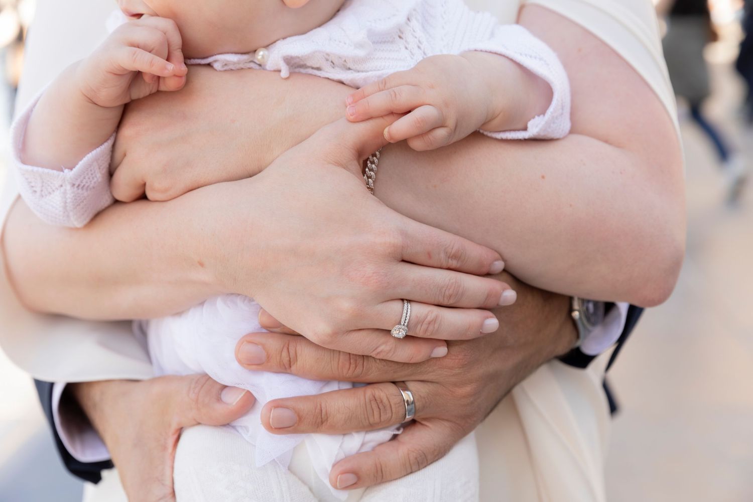 Close-up of parents hands holding a baby in white christening outfit, mother wearing diamond engagement ring.