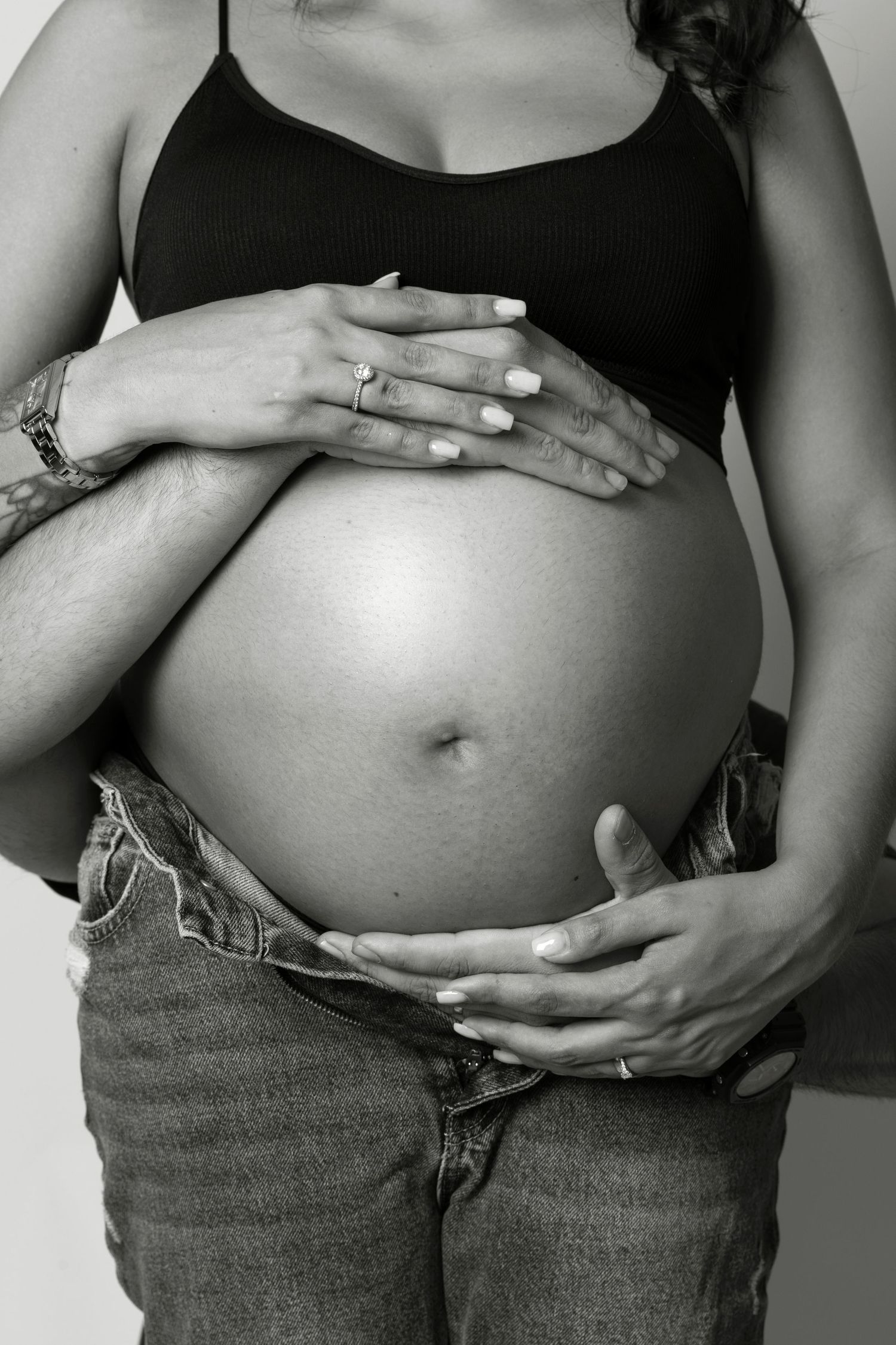 Black and white photo of a pregnant woman cradling her bare baby bump with a partner's arms wrapped around her.