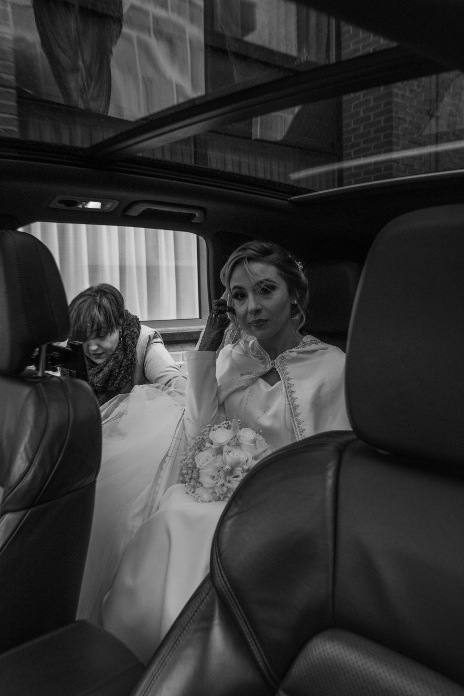Black and white photo of a bride in a wedding dress sitting in a luxury car, holding a bouquet, with a companion beside her.