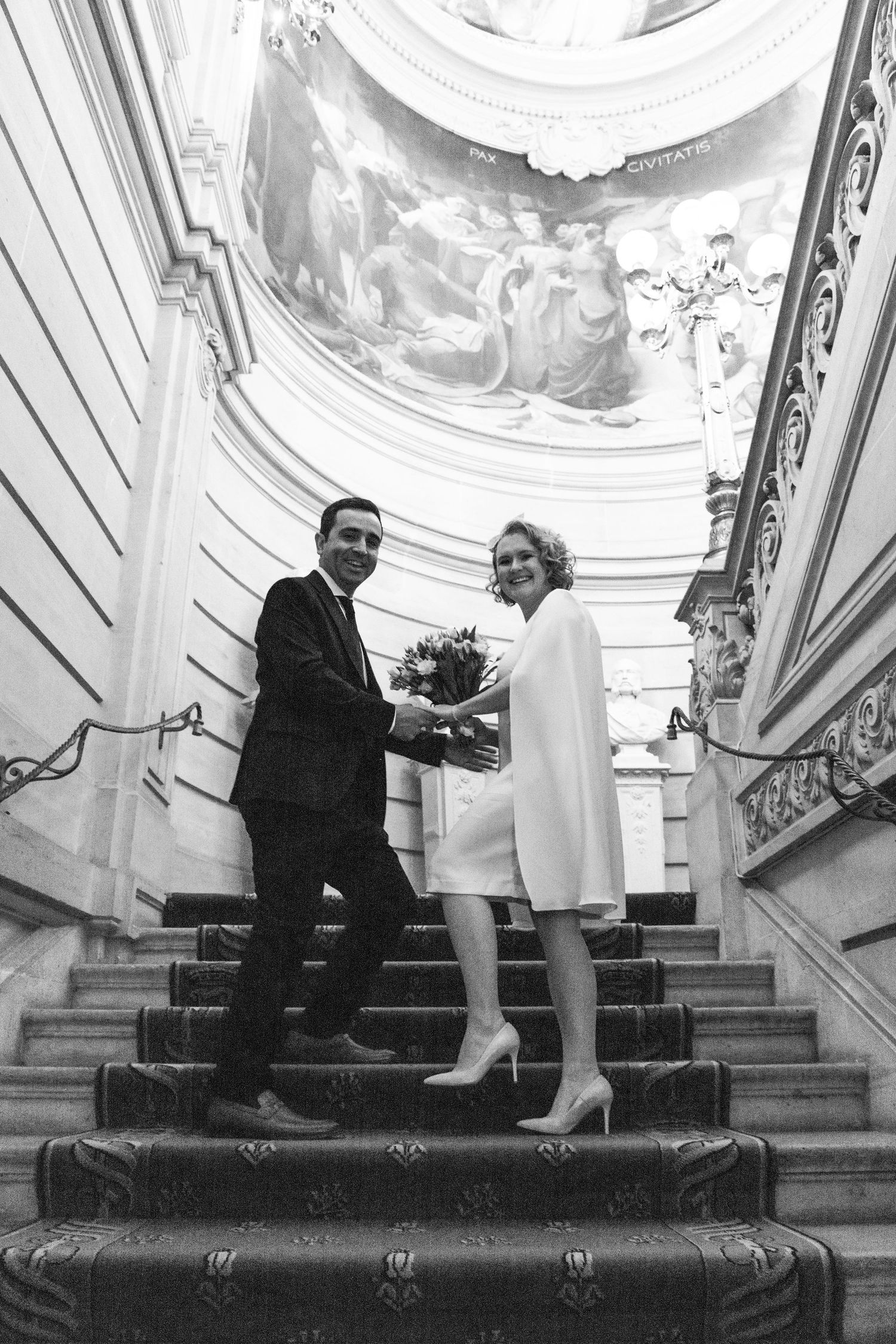 Elegant couple in wedding attire pose on grand staircase with ornate ceiling fresco in black and white.