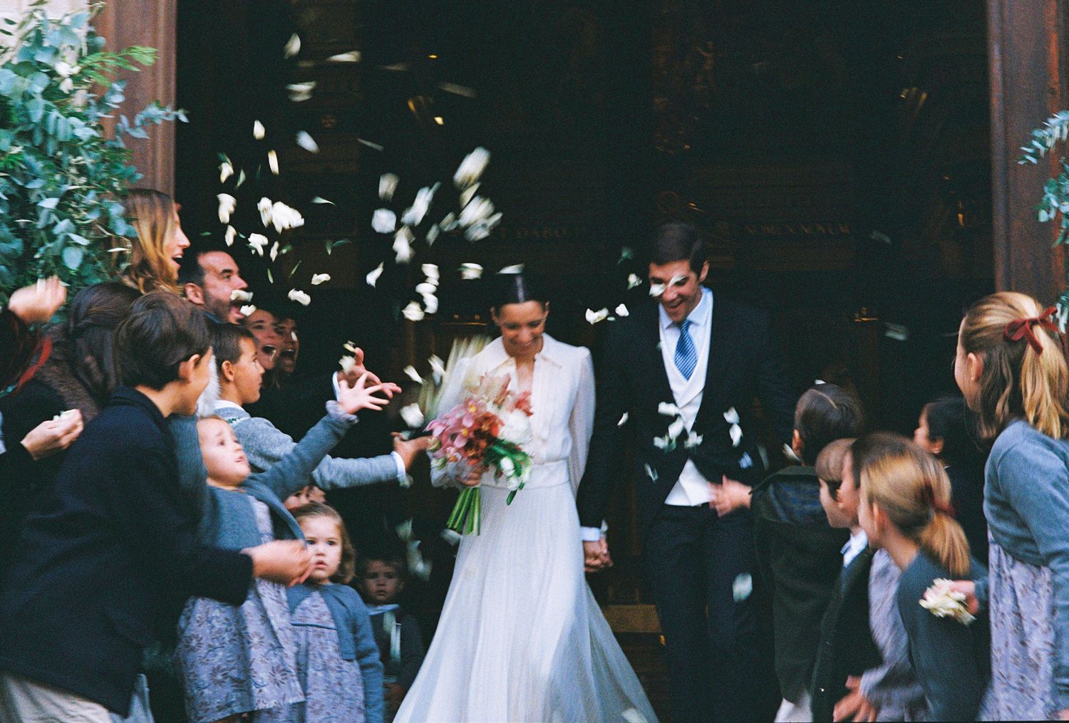 Bride and groom exit church as guests toss white flower petals in joyful wedding celebration.