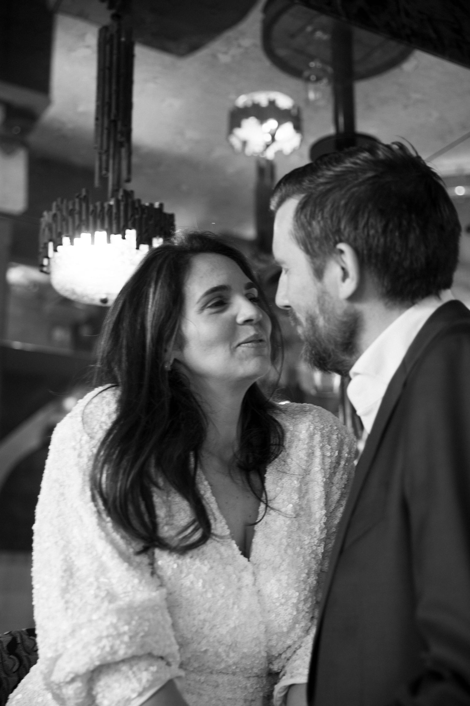 Black and white photo of a couple sharing a romantic moment at an elegant venue with chandeliers.