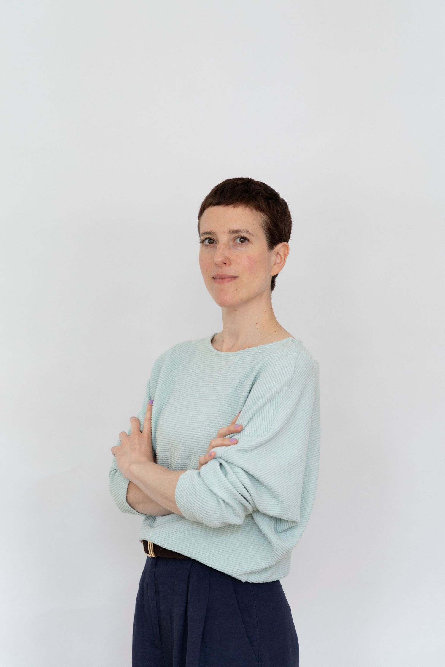 Woman with short hair in mint green sweatshirt and navy pants, arms crossed, posing against white background.