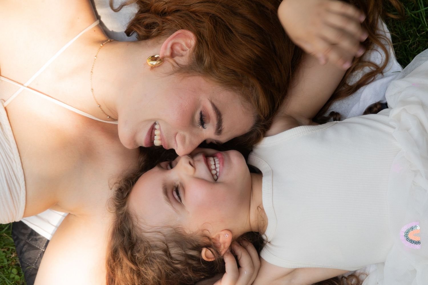 Two young women lying on grass, smiling and laughing together in a joyful, candid outdoor portrait.