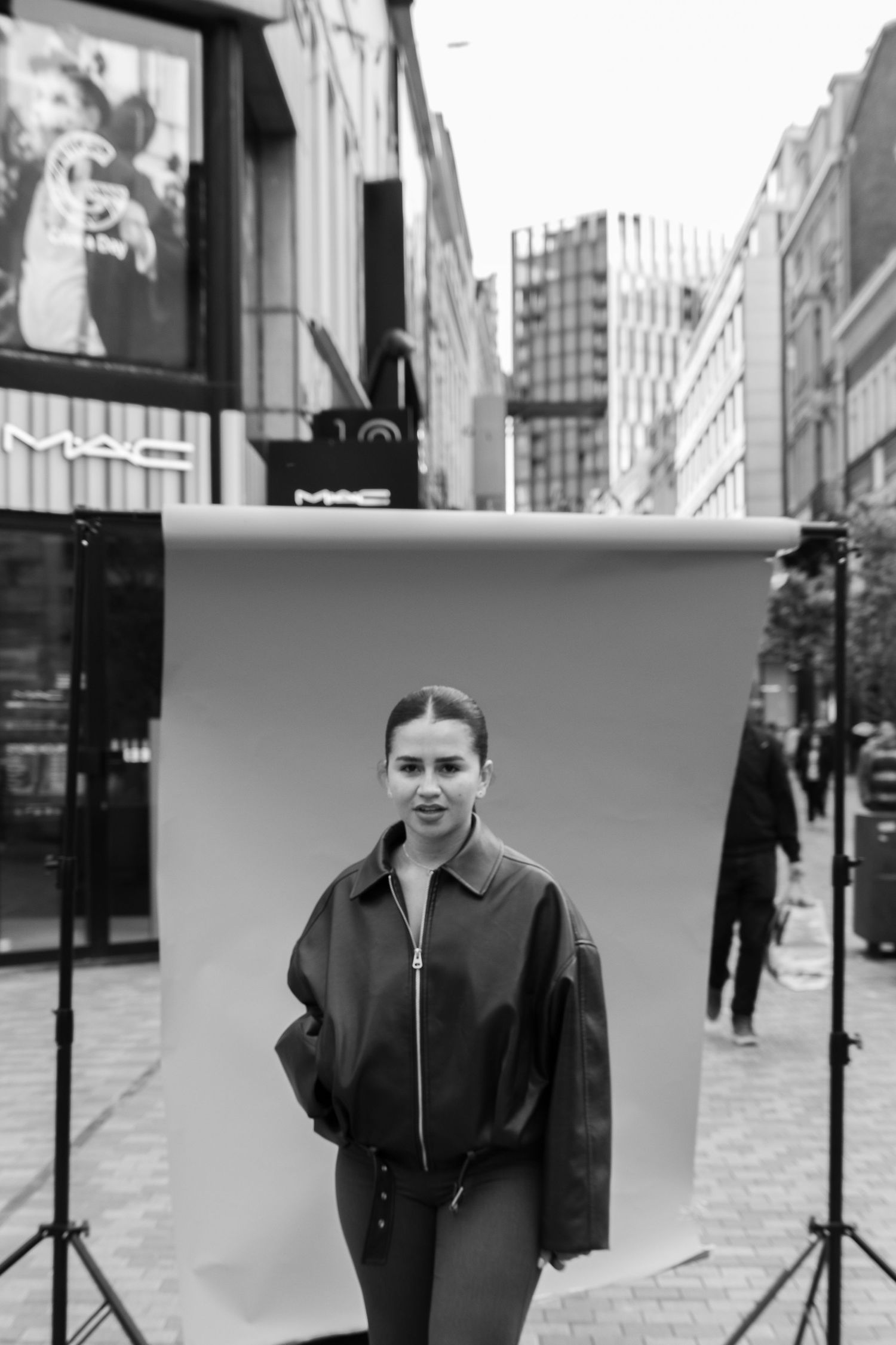 Black and white photo of a woman in a leather jacket posing in front of a backdrop on a city street near a MAC store.