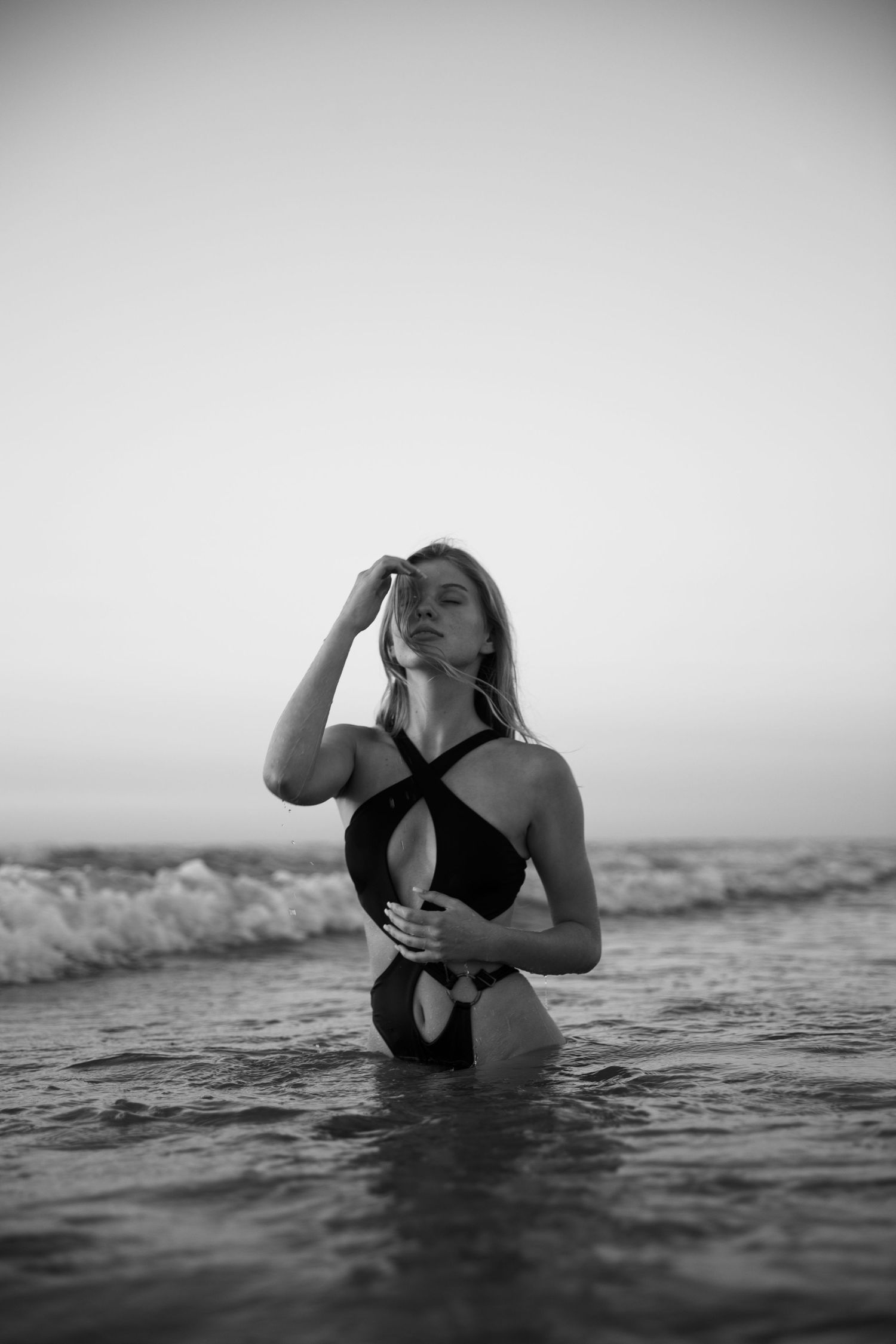 Black and white photo of a woman in a swimsuit standing in ocean waves at dusk. photography brussels