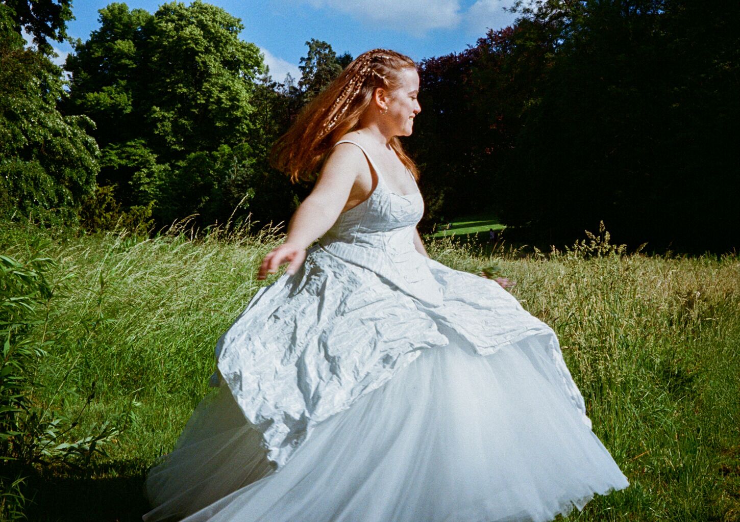 A woman in a flowing white wedding gown twirls gracefully in a lush green meadow surrounded by tall trees.
