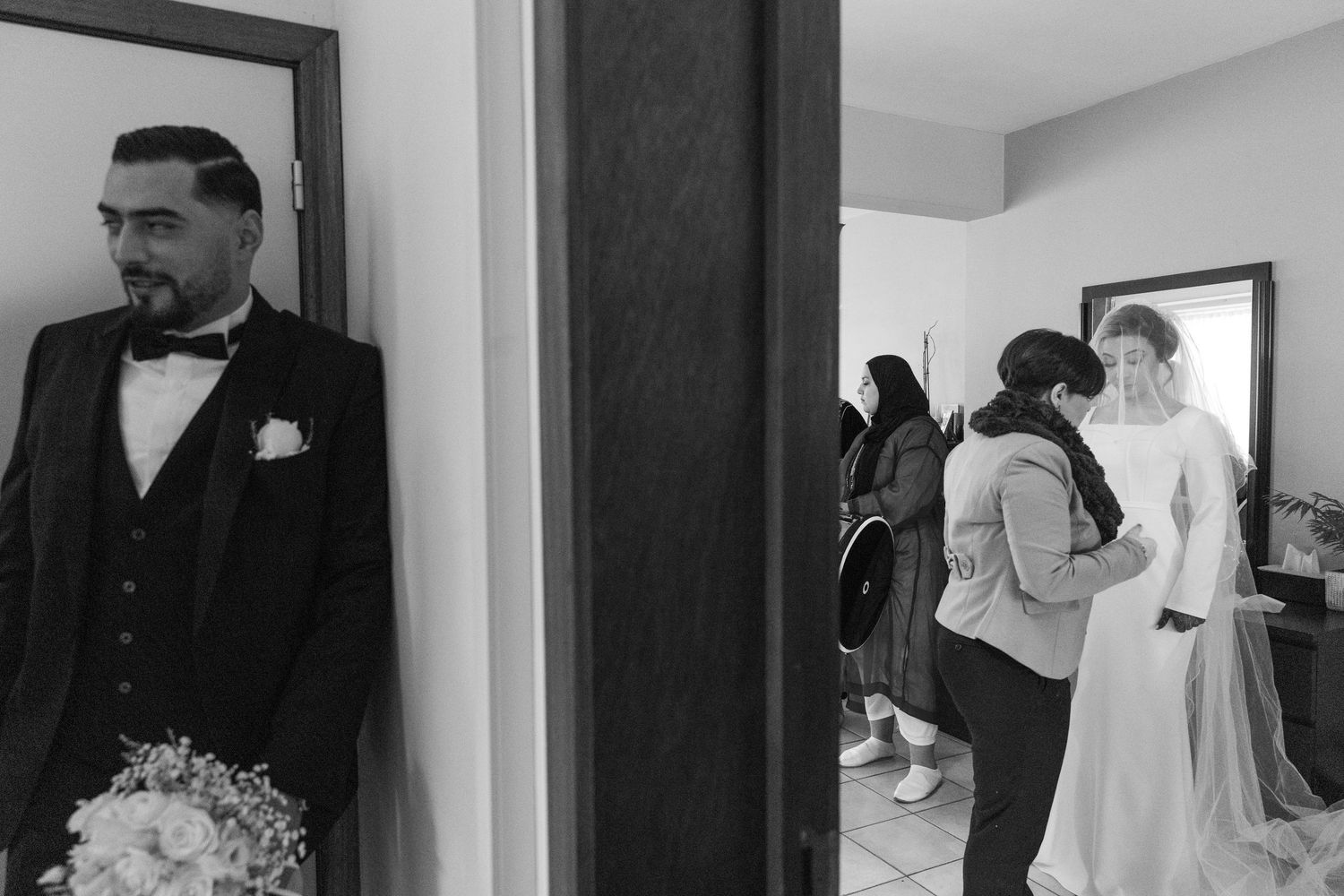 Black and white wedding photo of groom in tuxedo waiting while bride is assisted by attendants in separate room.