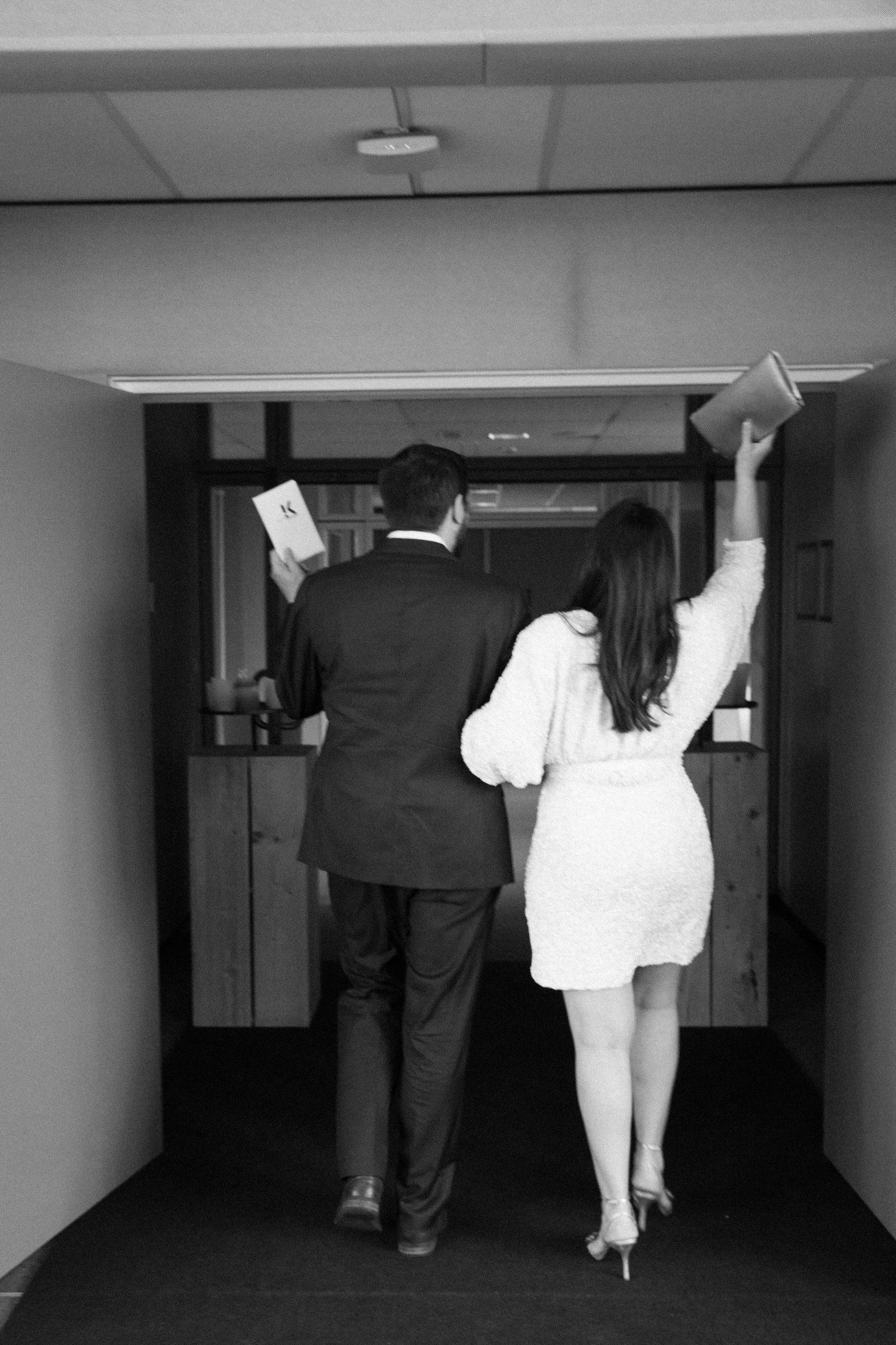 Black and white photo of a couple in formal attire walking down a hallway, woman raising a book in celebration.