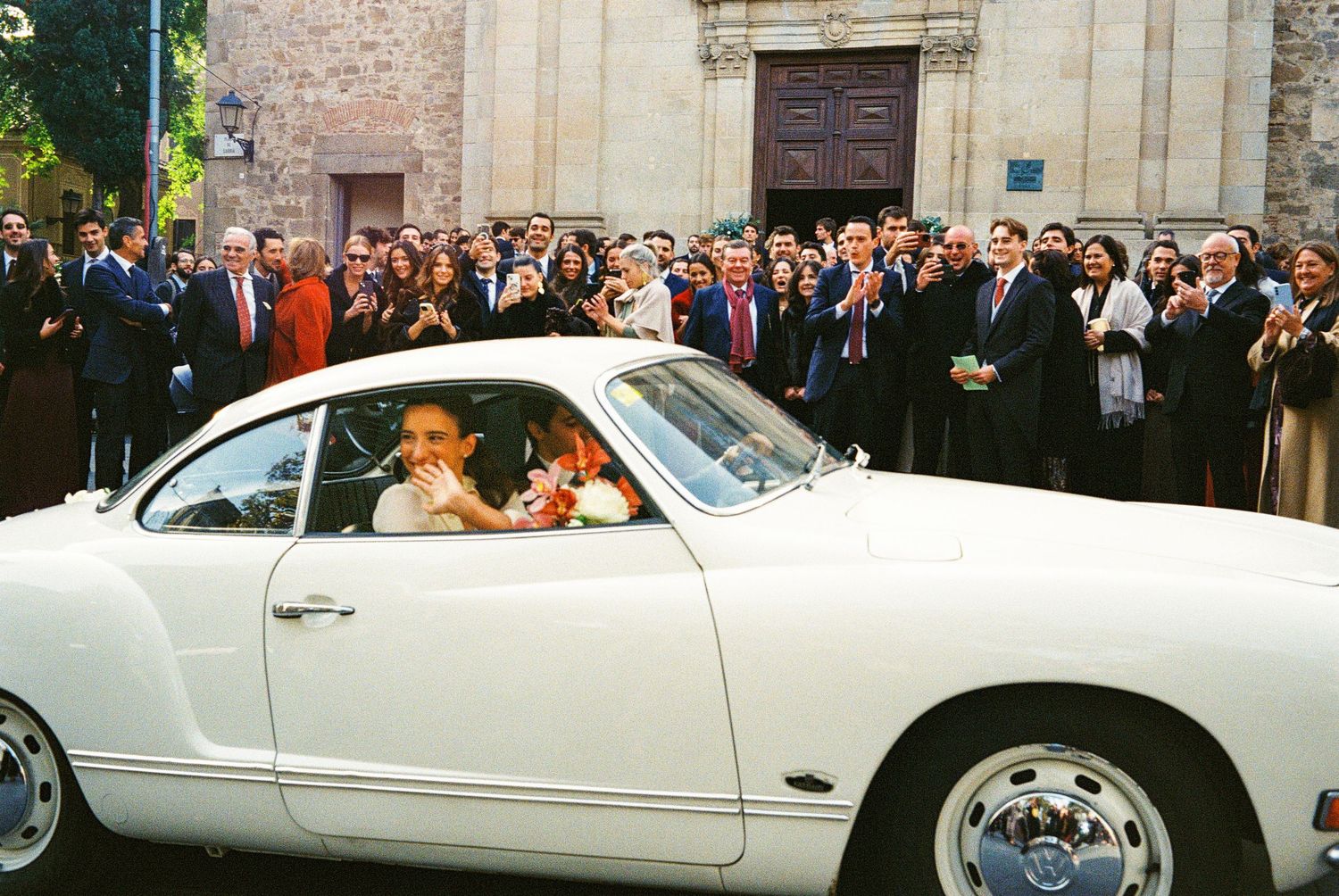 Bride and groom in a classic white Volkswagen Karmann Ghia surrounded by cheering wedding guests outside a church.