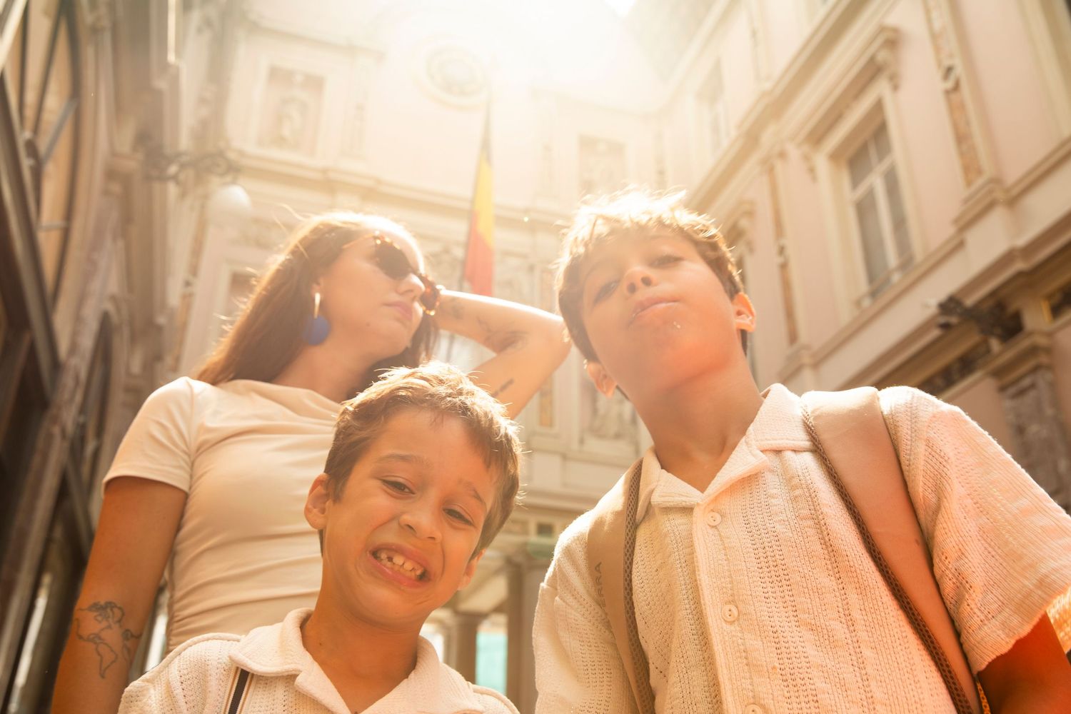 A mother and two young boys smile outdoors in bright sunlight near a historic building on a sunny day.
