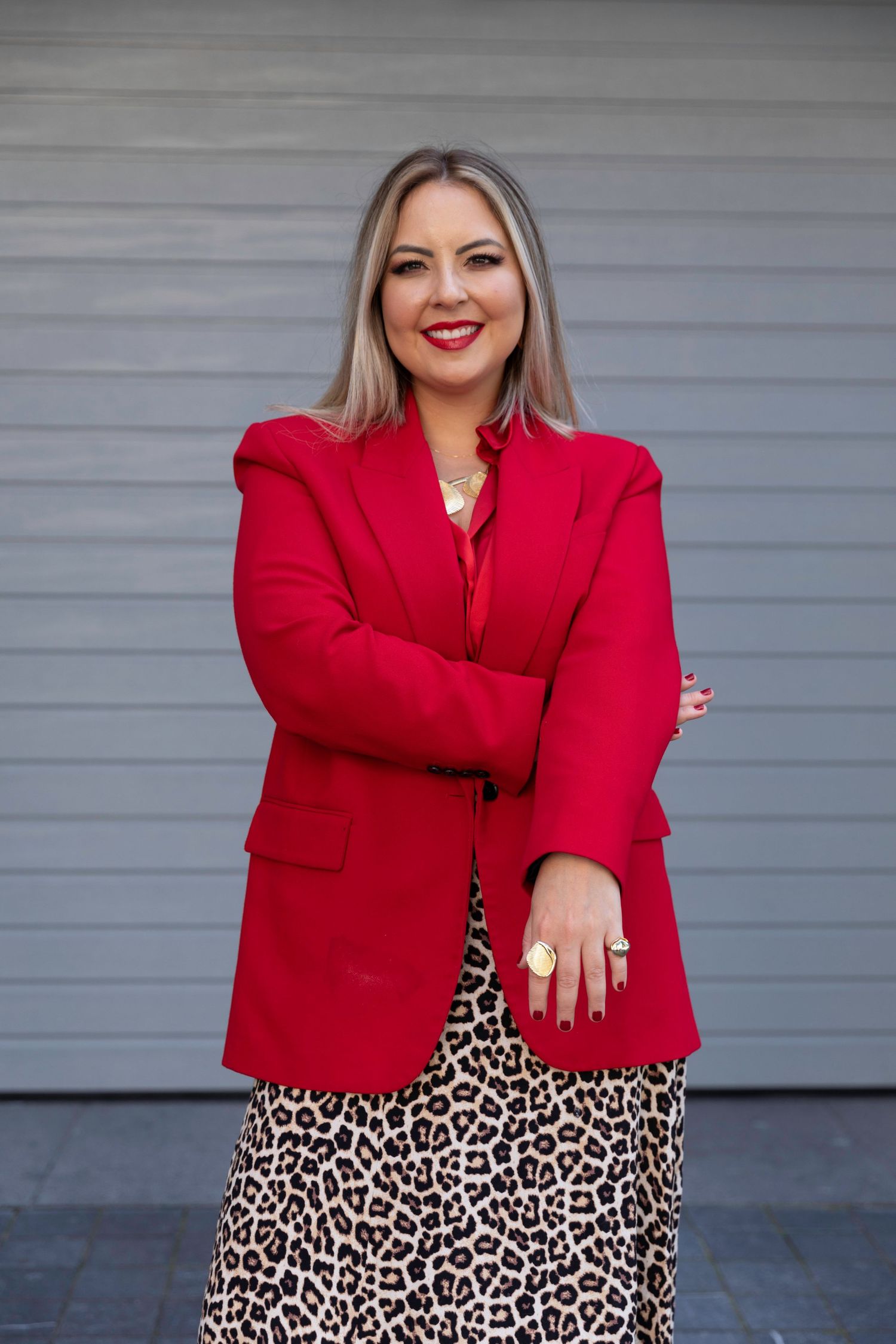 Woman in red blazer and leopard print skirt smiling confidently with arms crossed against gray wall.