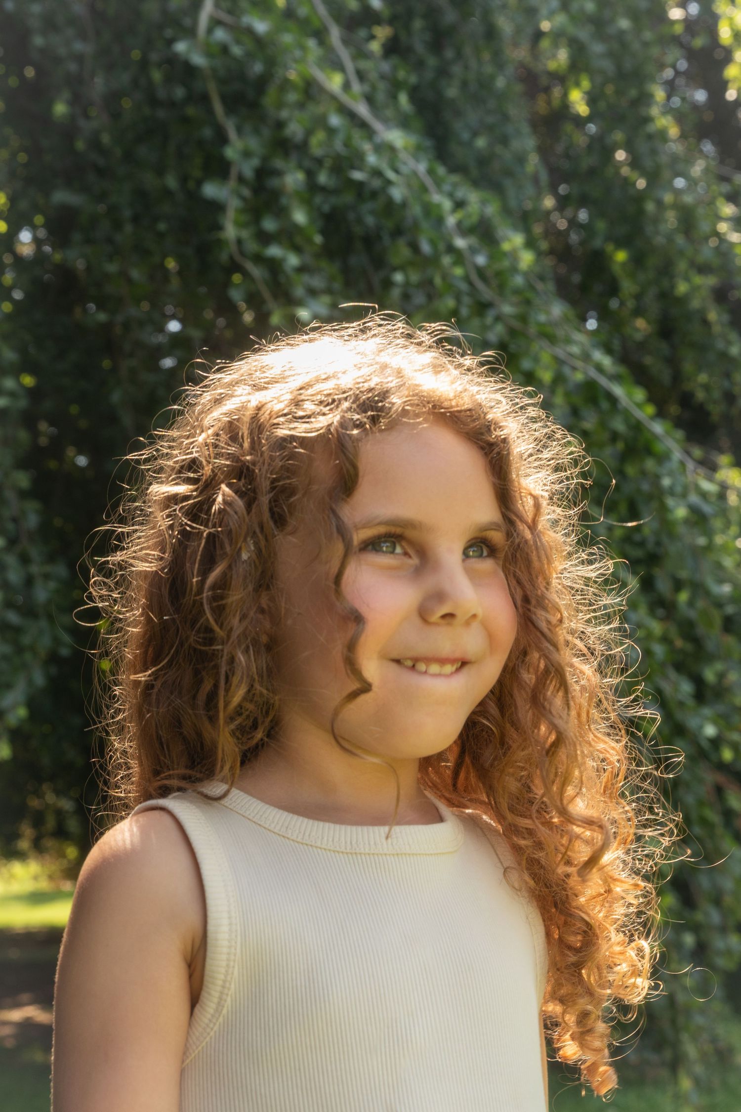 Smiling young girl with curly auburn hair wearing a cream sleeveless top, standing outdoors with green trees in background.