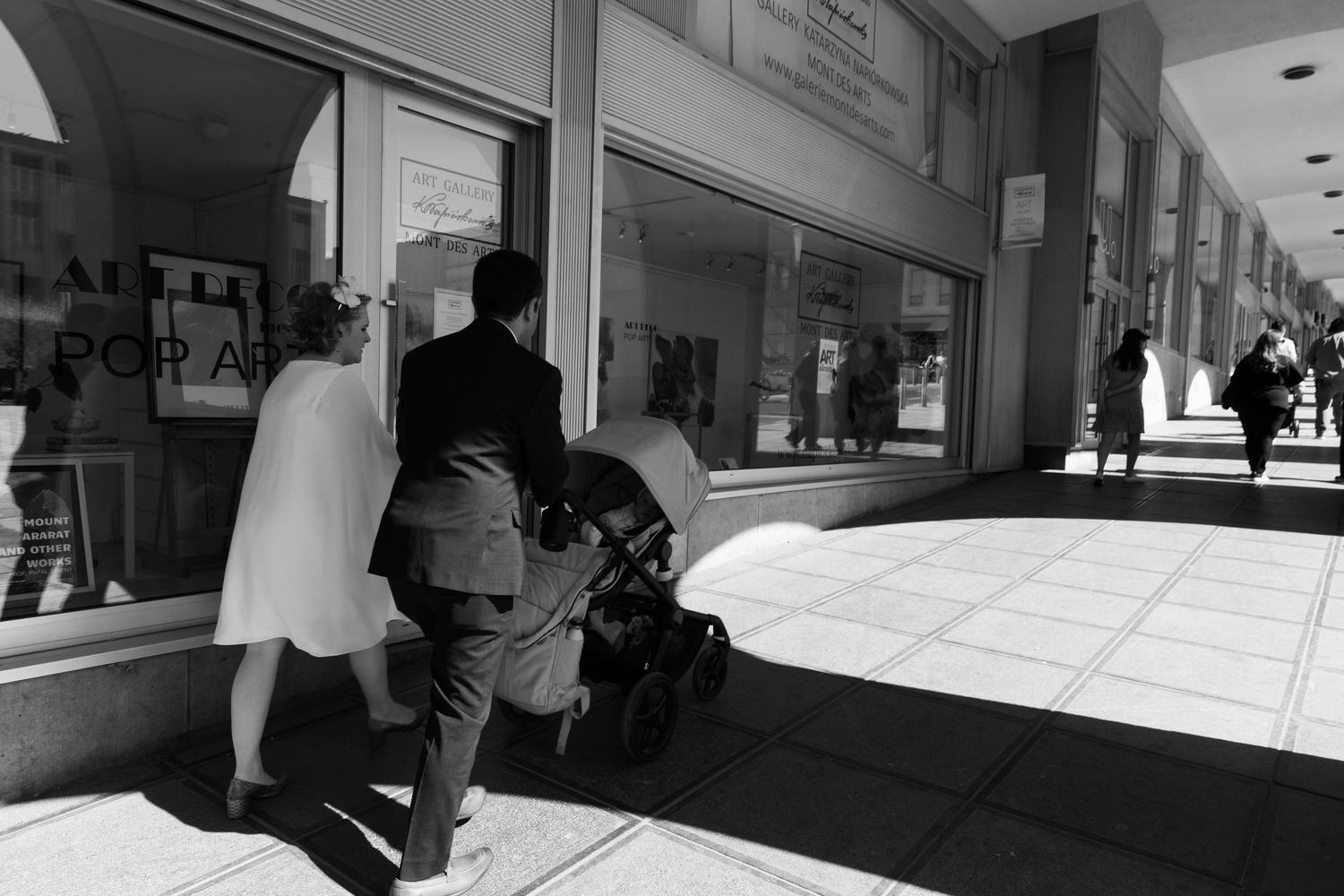 Black and white photo of a couple in wedding attire pushing a stroller along a sunny urban sidewalk.