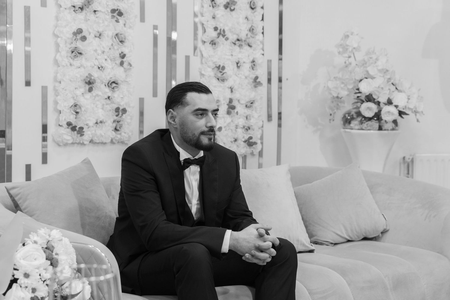 Elegant man in black tuxedo with bow tie sitting on sofa at formal event.